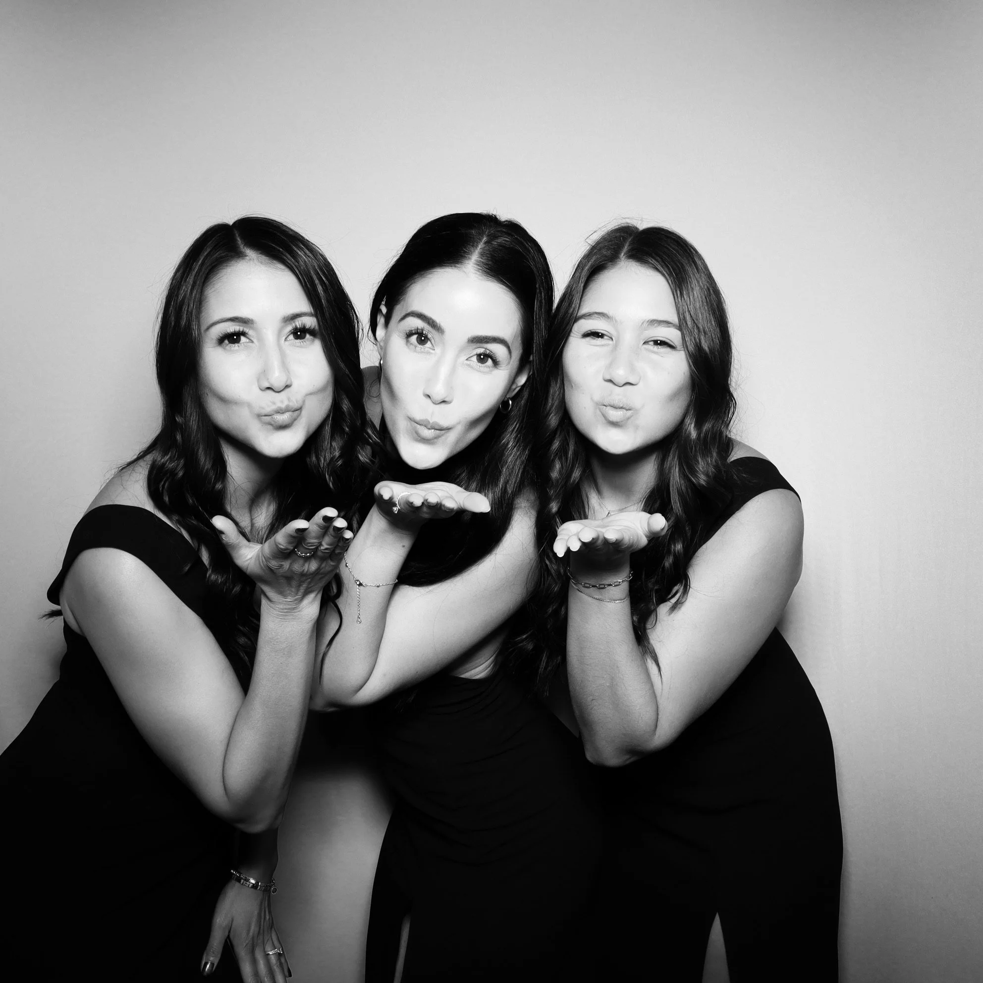 Three women with long dark hair blowing kisses to the camera, smiling, in a black and white photo.