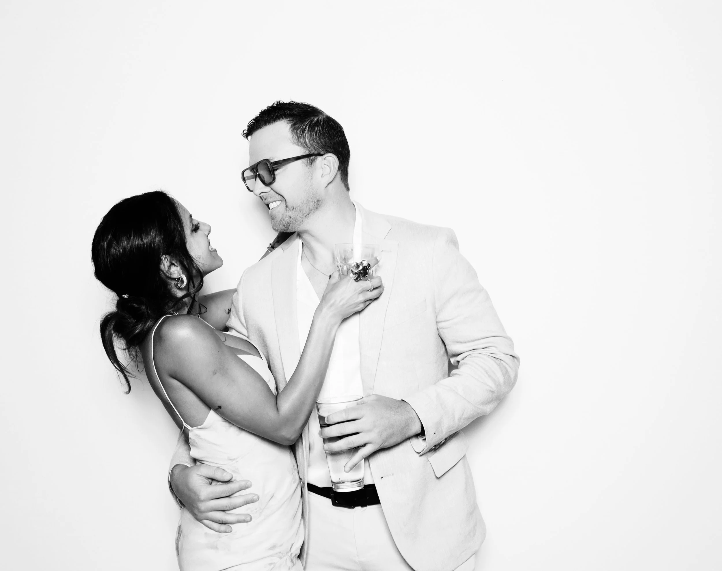 A man and woman smiling at each other kissing while holding drinks in front of a plain white background.