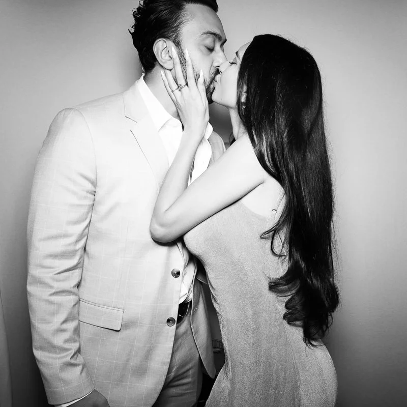 A couple sharing a kiss, the man in a light suit and the woman in a sleeveless dress, with her hand on his face.