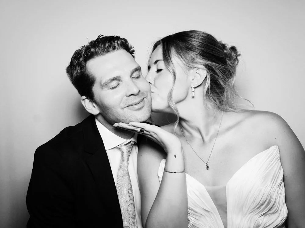 A woman in a wedding dress is kissing a man in a suit on the cheek, with her hand gently touching his chin.