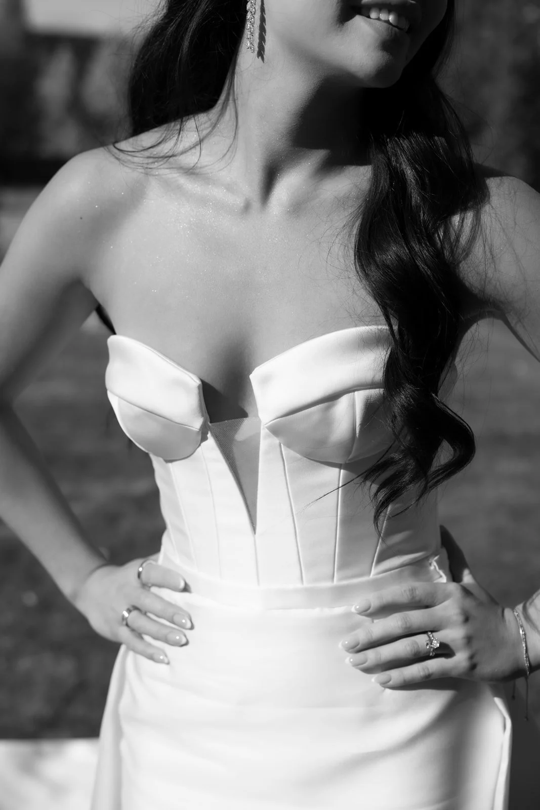 A woman wearing a strapless dress with a fitted bodice and long, wavy dark hair. She has her hands on her hips, showing rings and bracelets, and is smiling.