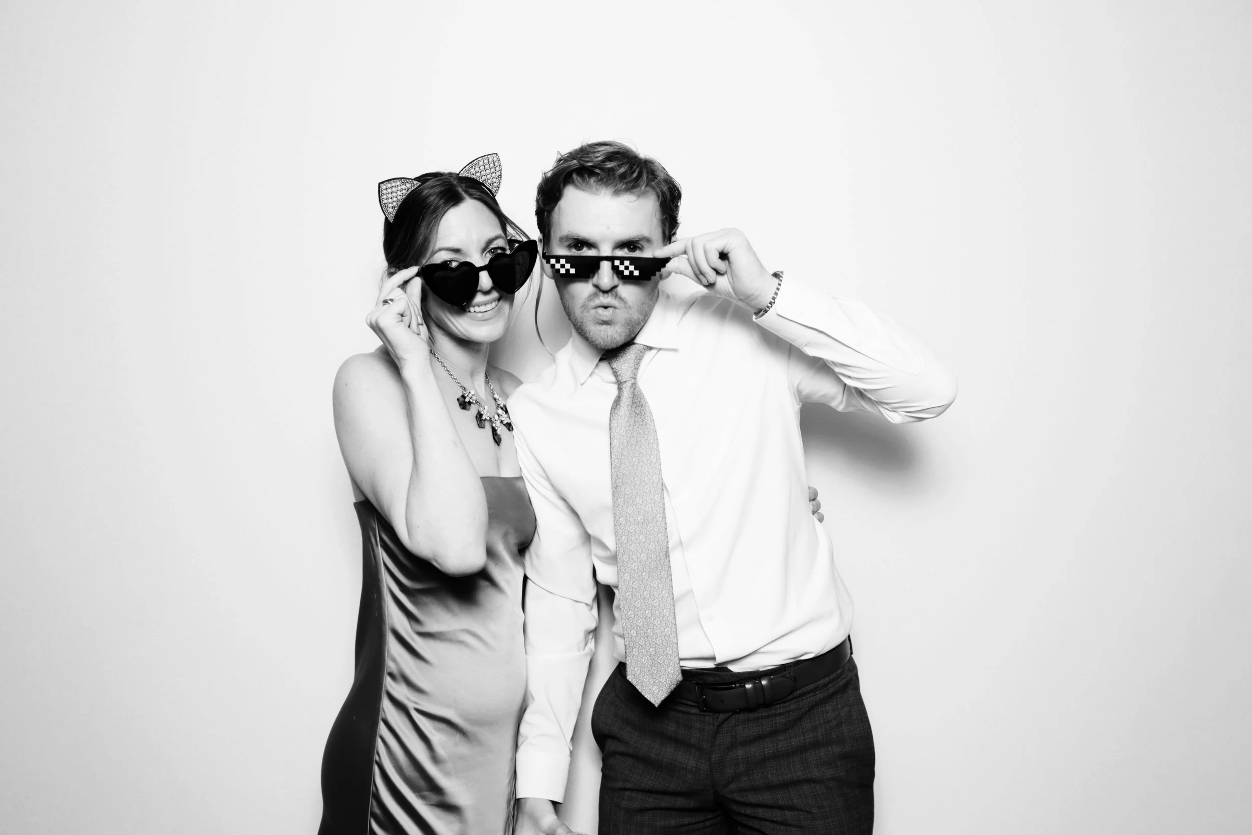 A young woman and man wearing sunglasses and party accessories posing for a photo in front of a plain white background.