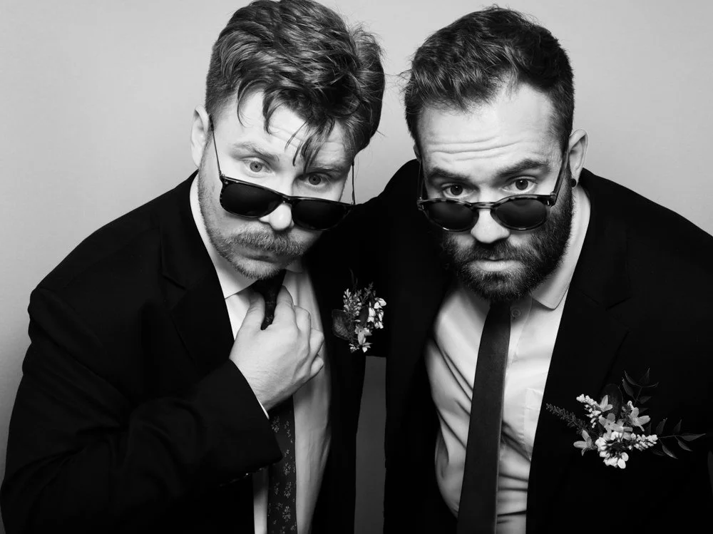 Two men in suits wearing sunglasses, looking directly at the camera, with serious expressions, against a neutral background.