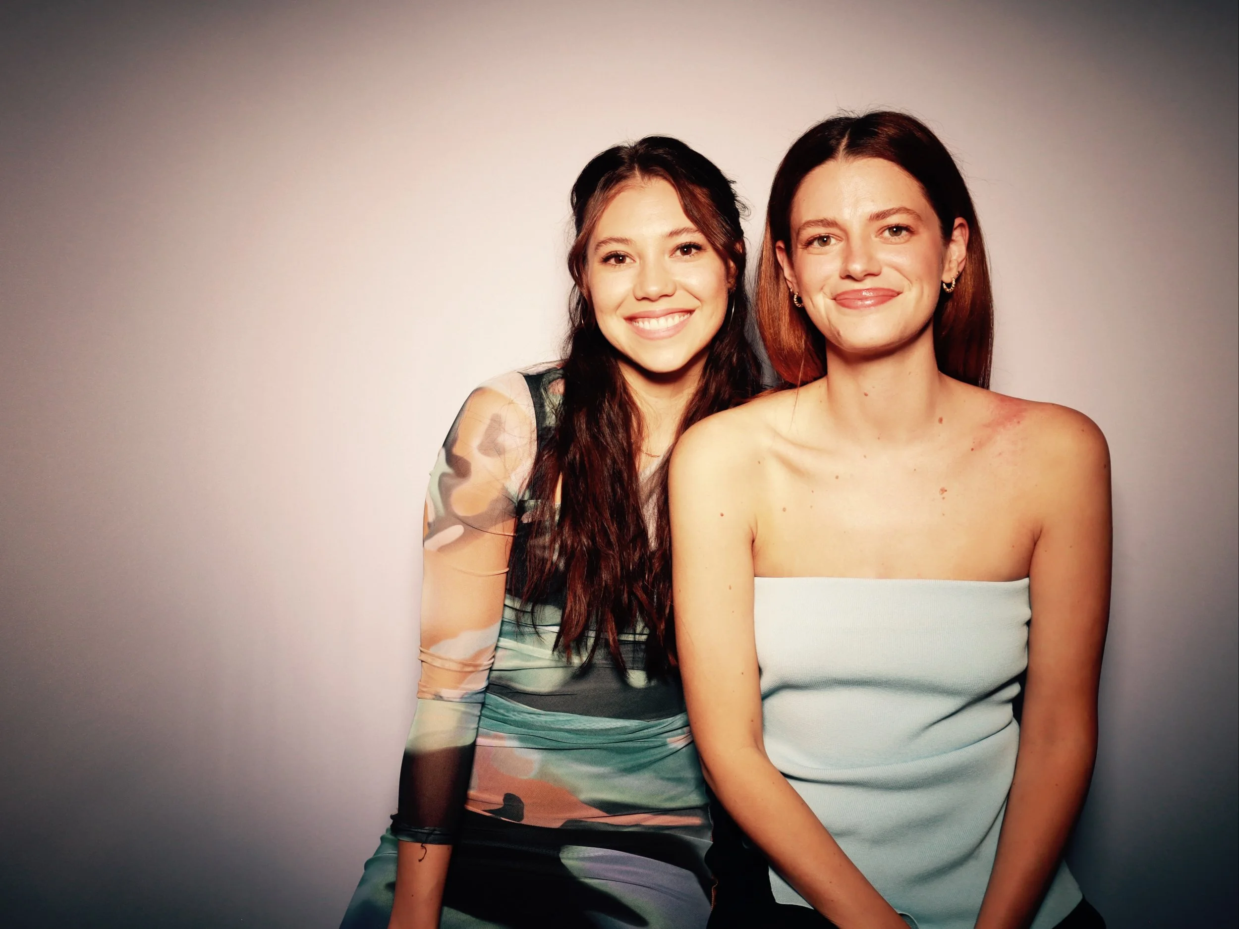Two women smiling, one with long dark hair and patterned dress, the other with shoulder-length brown hair and strapless white top, sitting close together against a plain background.