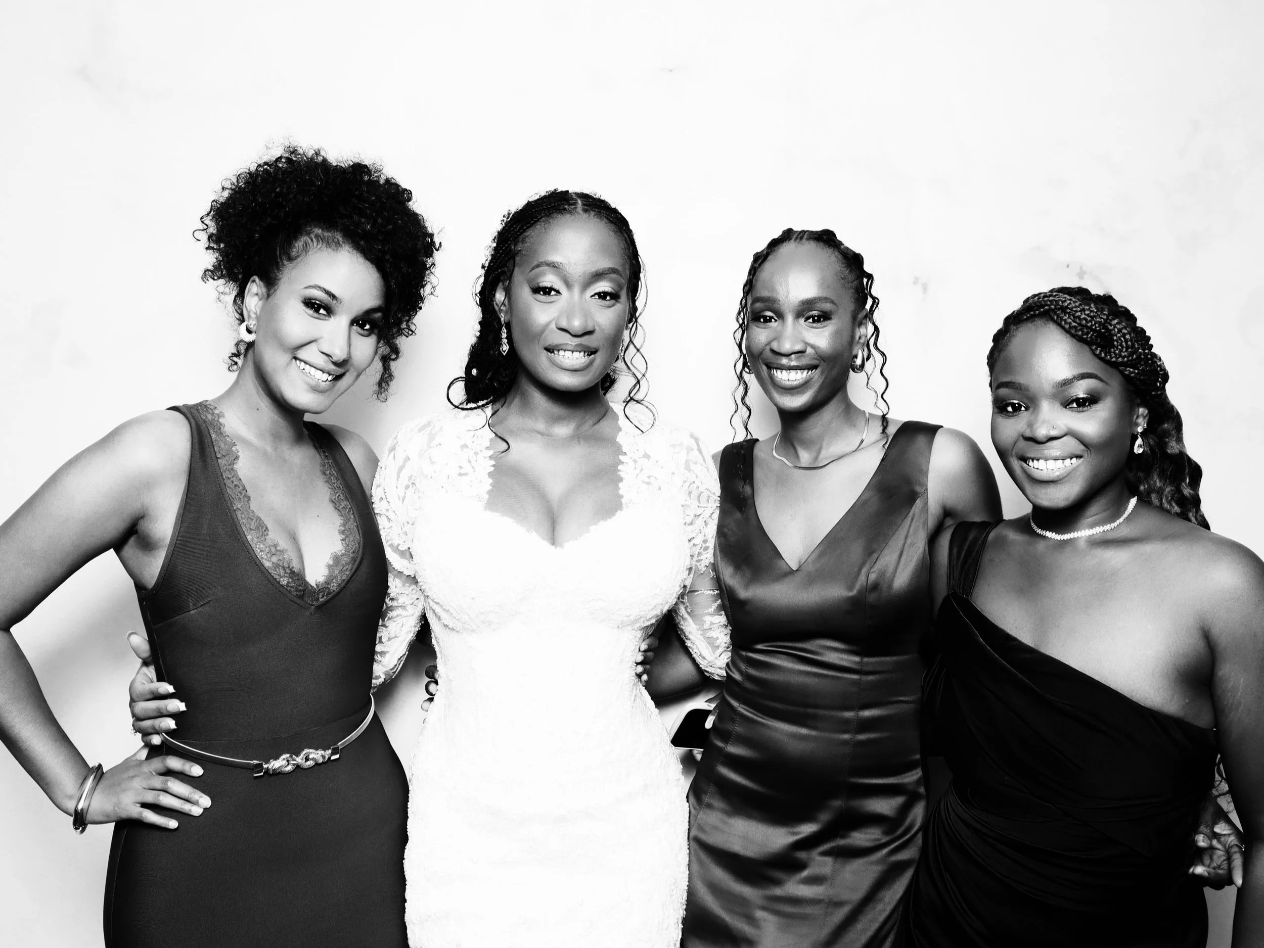 Four women in elegant dresses smiling and posing together in a black and white photo.
