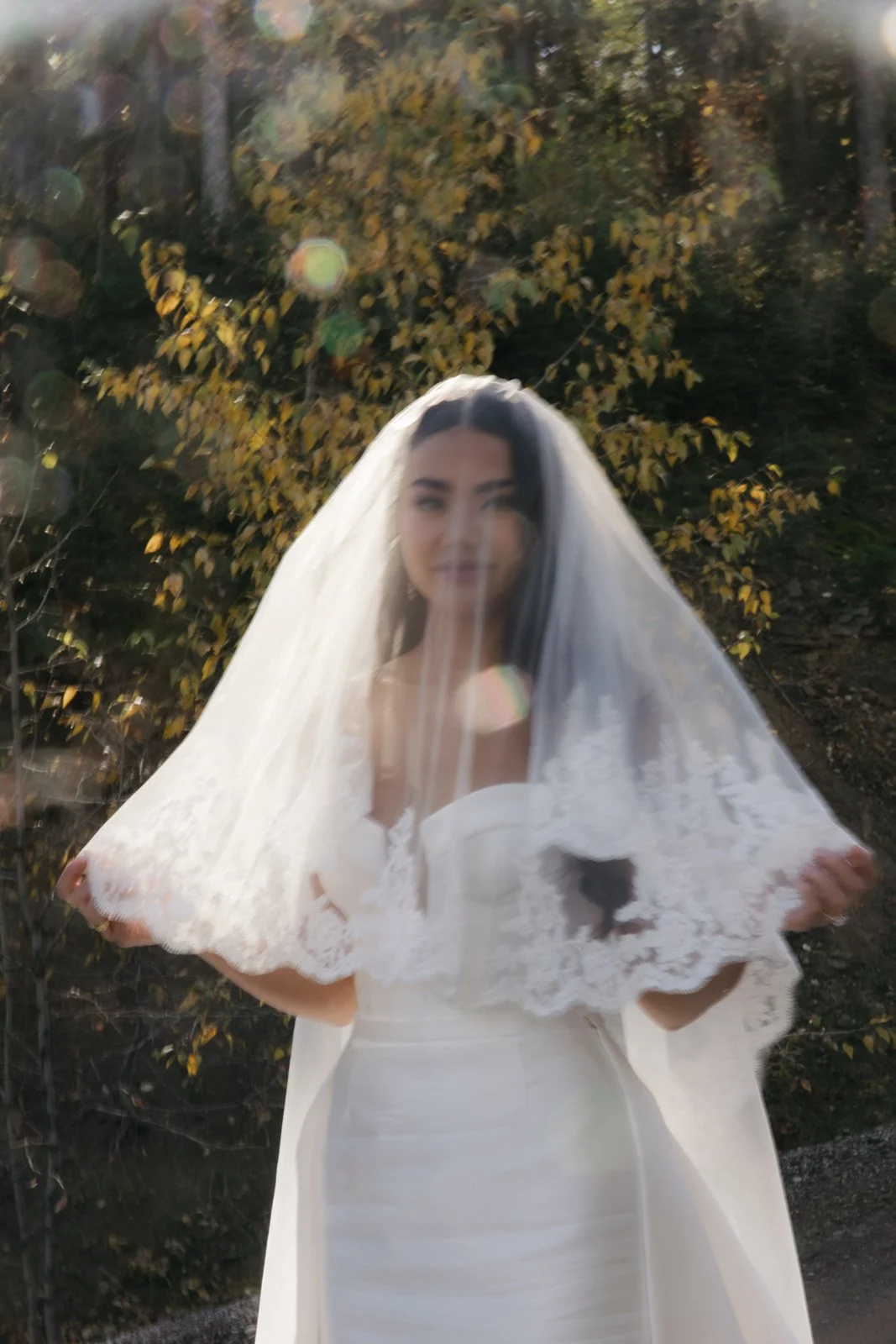 A woman wearing a wedding dress and veil holds up the veil in front of her face, standing outdoors with trees and sunlight in the background.