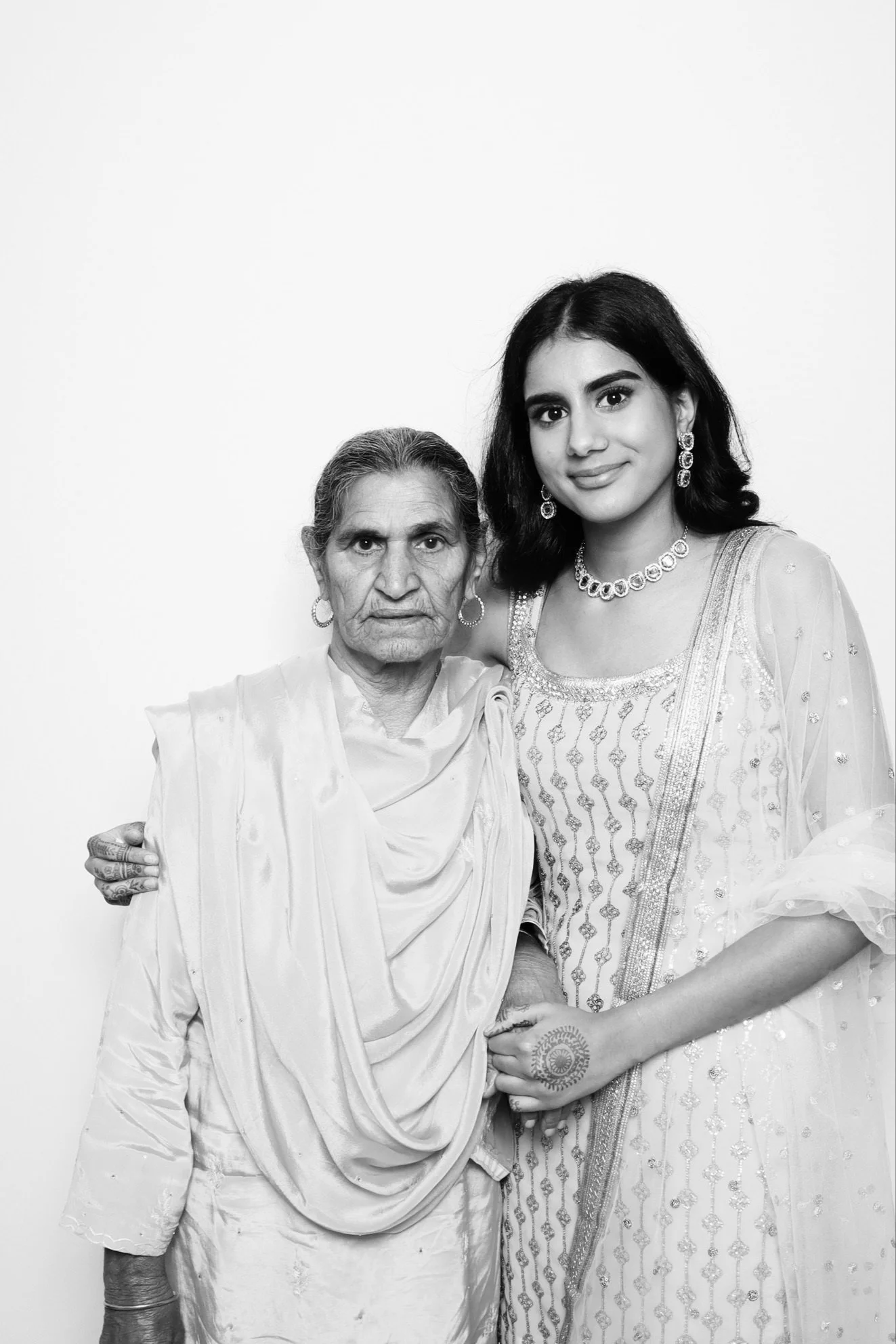 A black-and-white photo of an elderly woman and a young woman standing close together, both dressed in traditional Indian attire with jewelry, peaceful expressions, and a plain white background.