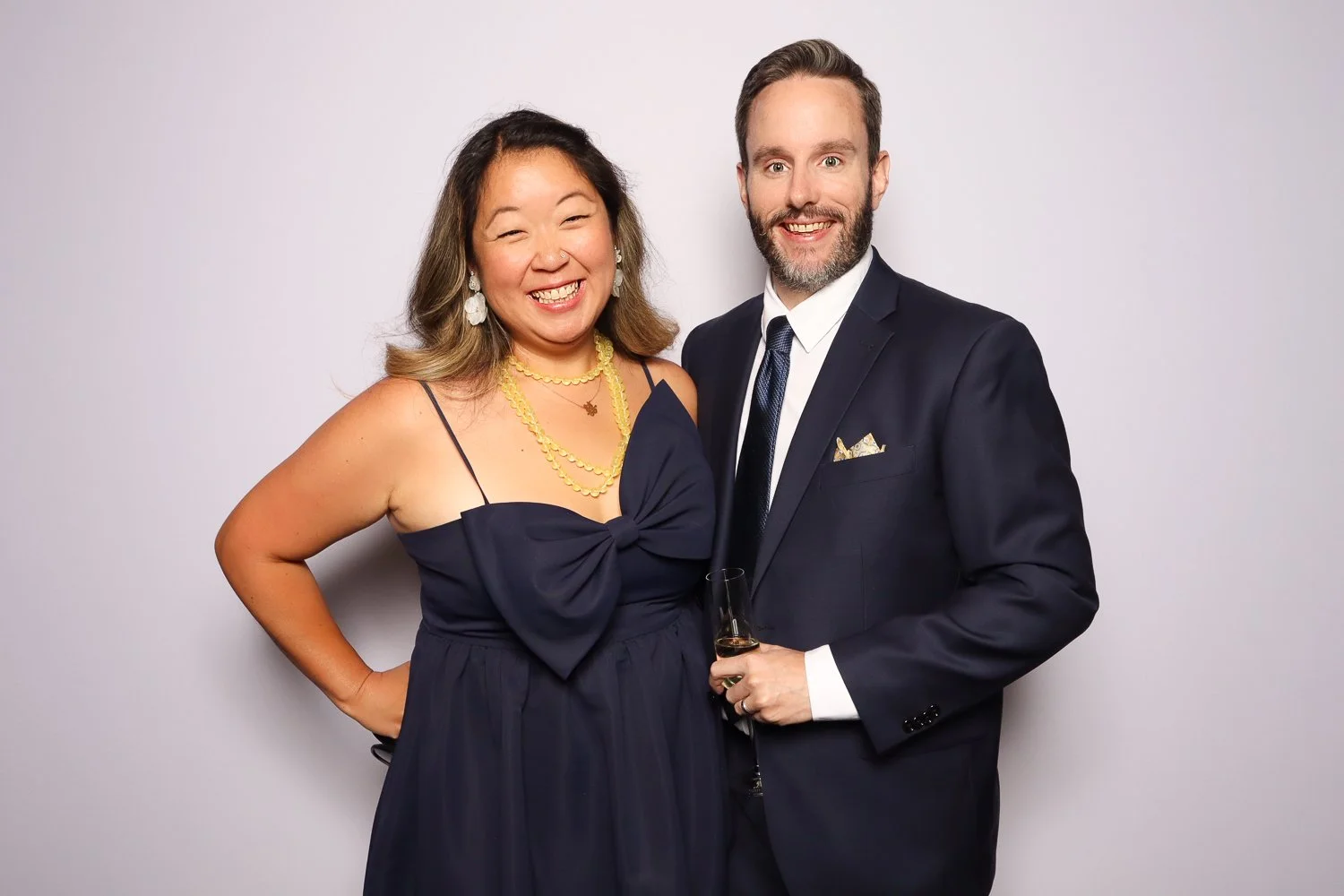 A smiling woman in a dark strap dress with a large bow, wearing yellow jewelry, and a man in a dark suit with a white shirt and tie, holding a glass, posing together against a plain light-colored background.