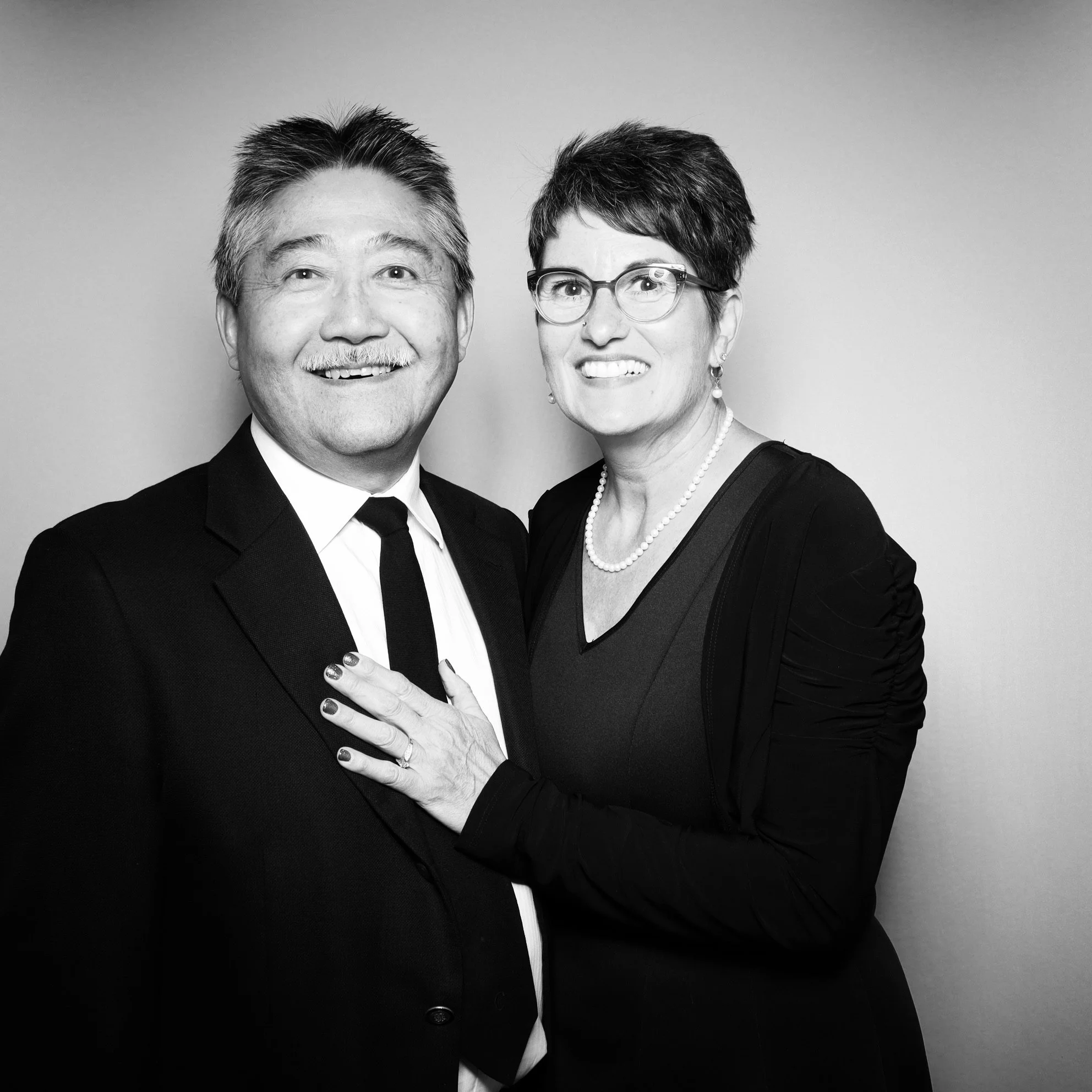 Black and white photo of a smiling middle-aged man in a suit and tie alongside a woman with short hair, glasses, pearl necklace, and black dress, standing close together against a plain background.