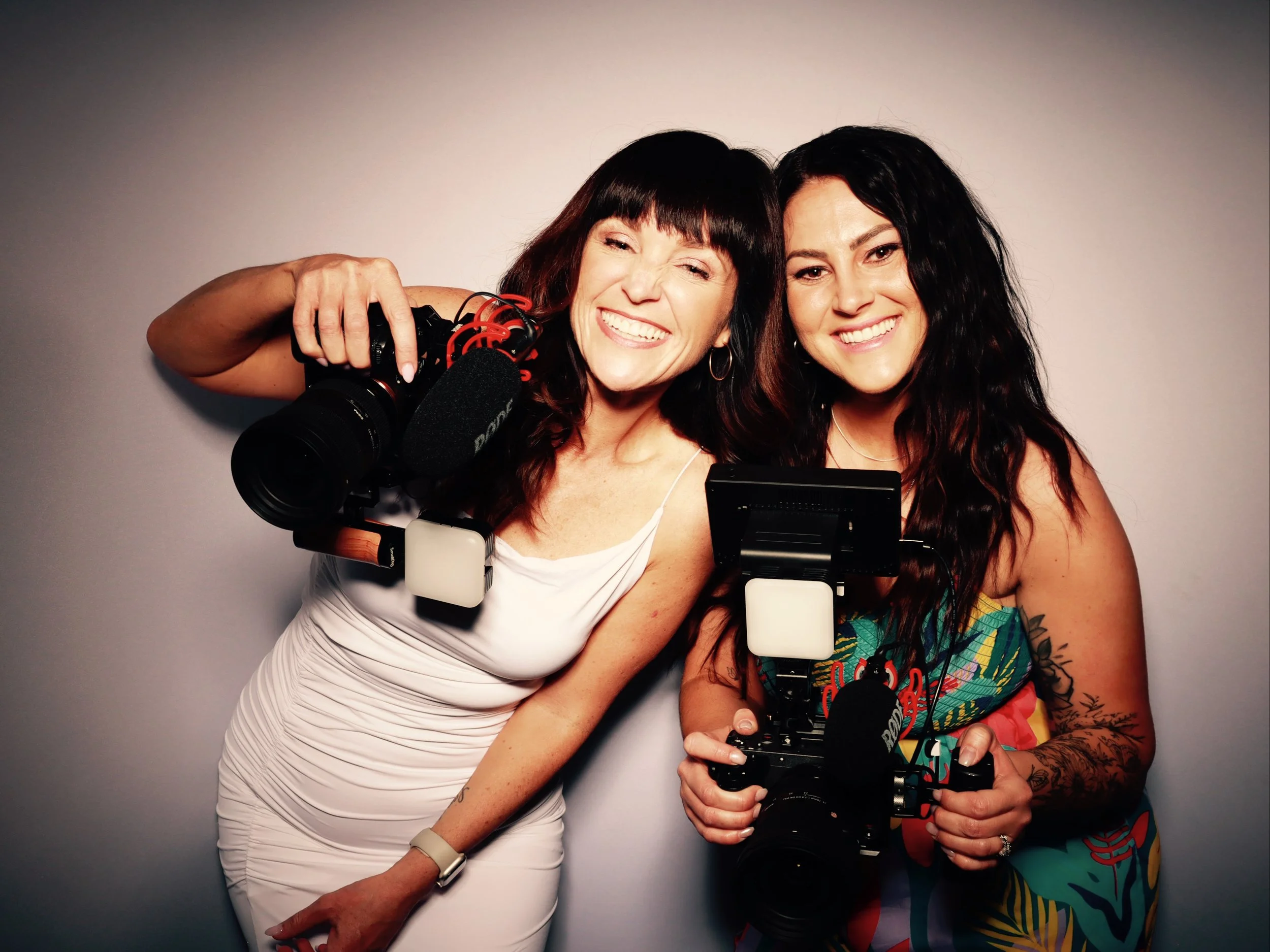 Two women smiling and holding video cameras, standing close together against a plain light gray background.
