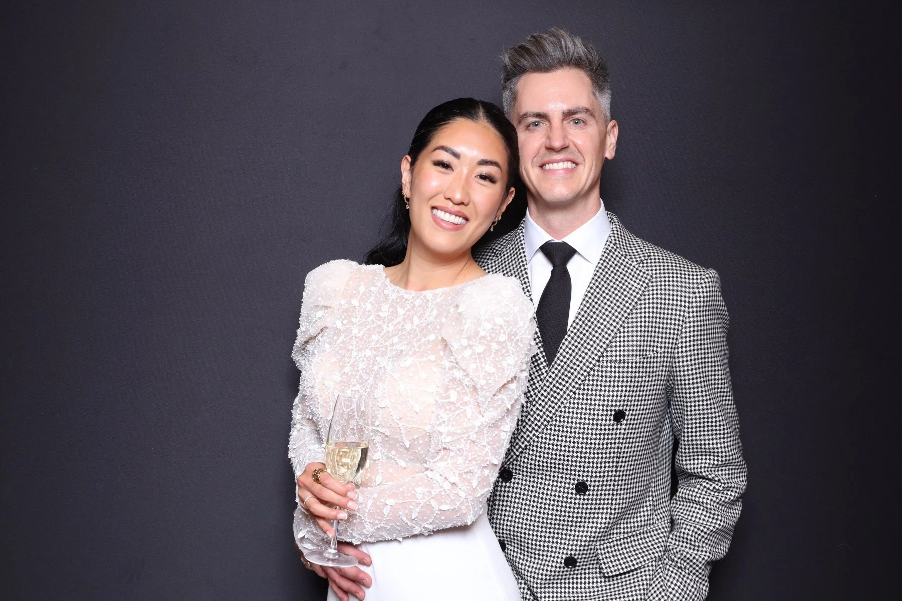 A smiling woman in a white, textured dress holding a glass of champagne, standing next to a smiling man in a black-and-white checkered suit with a black tie. They are posing together against a dark background.