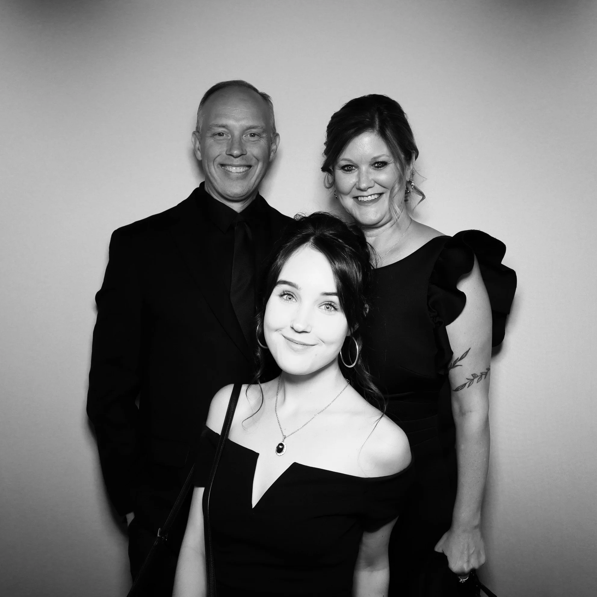 Black and white photo of three smiling people in formal attire, a young woman in the foreground and a man and woman behind her, against a plain background.