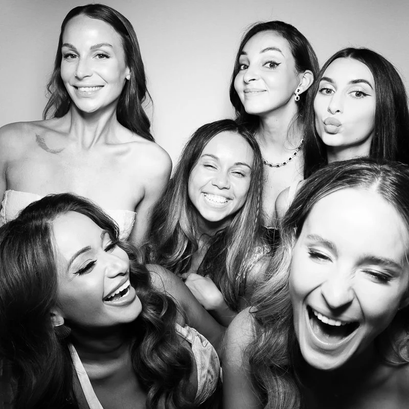 Six women smiling and laughing in a group photo, some with playful expressions, in black and white.