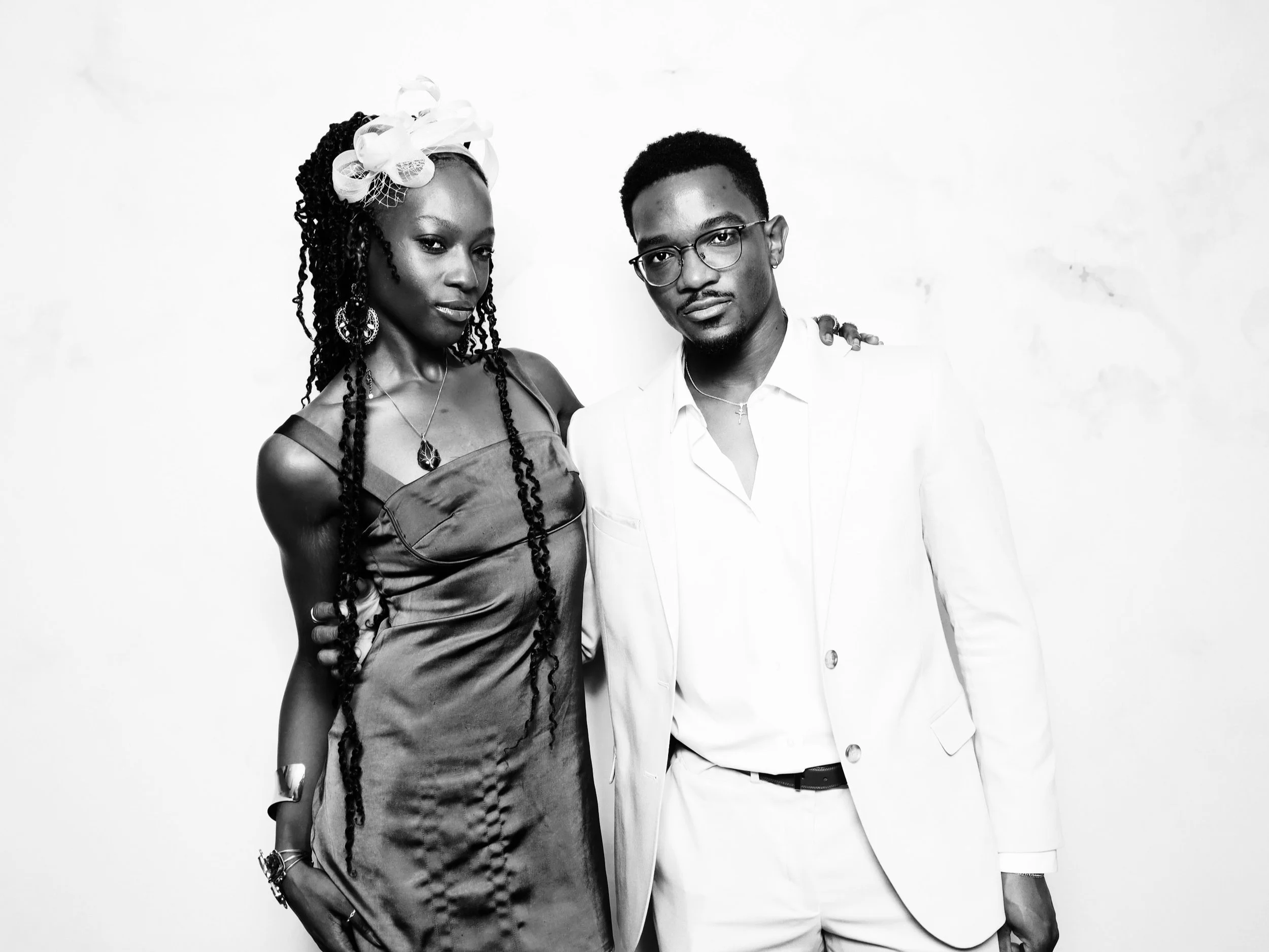 A black and white photograph of a woman in a satin dress with jewelry and a man in a white suit with glasses, posing against a plain white background.