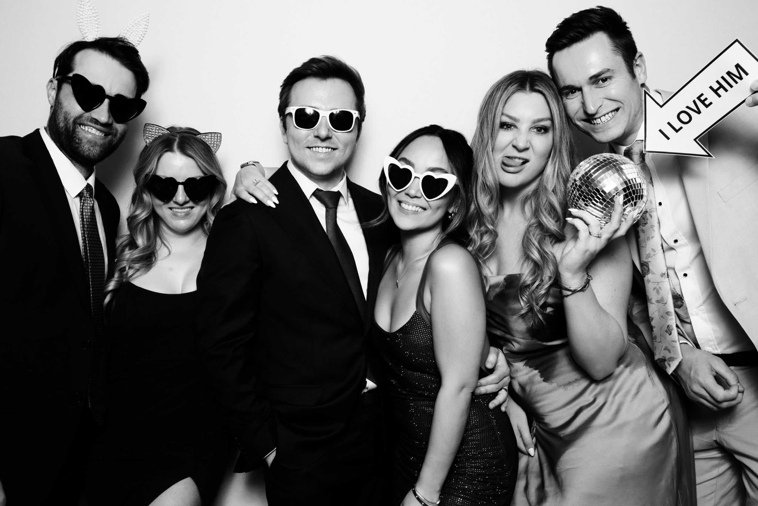 Black and white photo of seven young adults at a party, wearing sunglasses, with some making playful faces or poses, one holding a disco ball and a sign that says 'I LOVE HIM'.