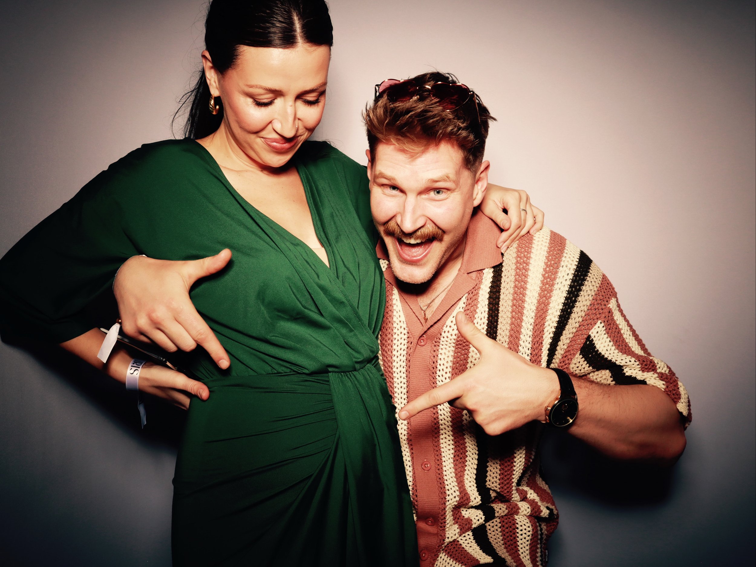 A smiling woman in a green dress with a pregnancy bump, and a man with a mustache and beard in a striped shirt, pointing at her belly.