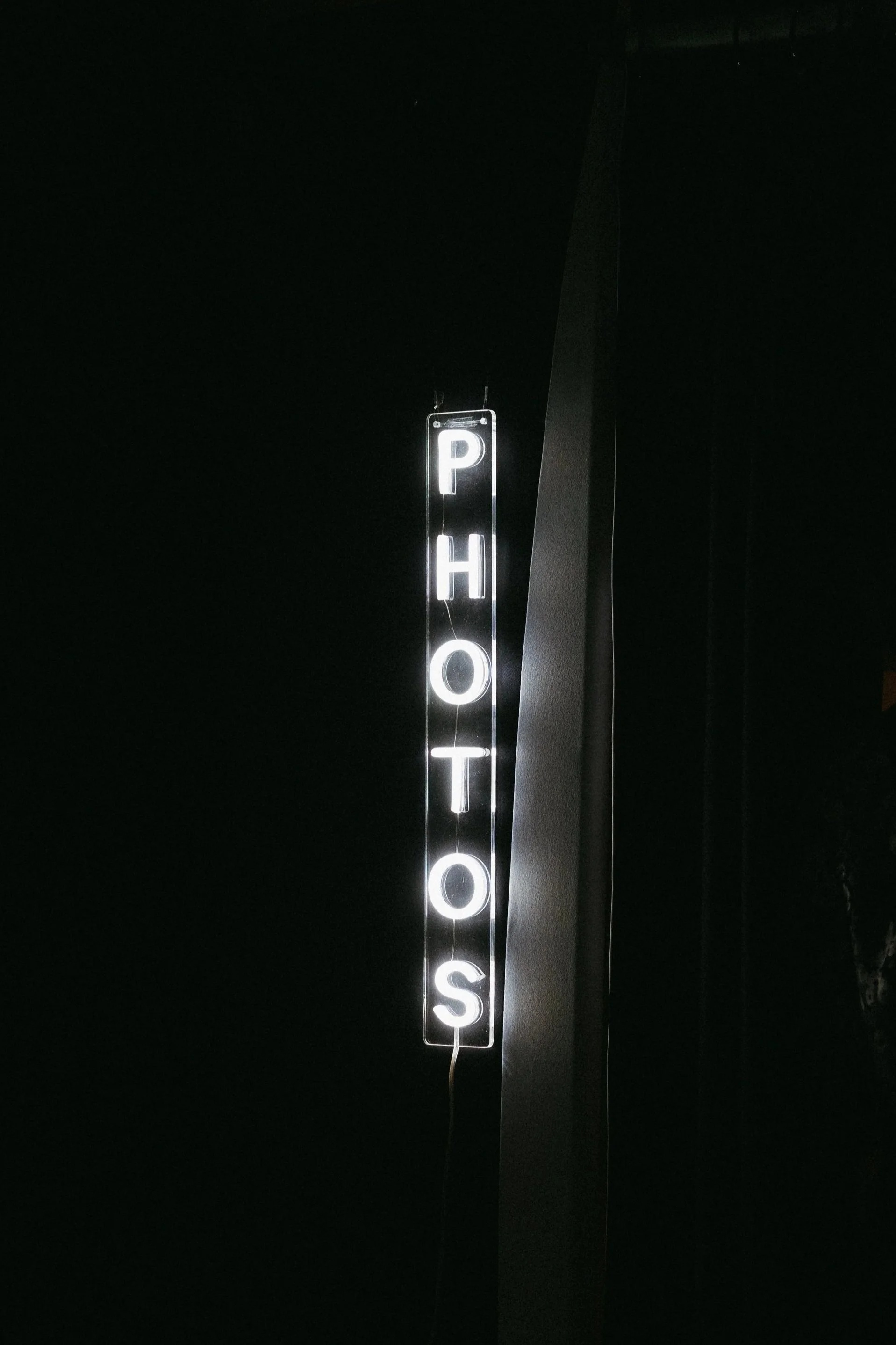 Illuminated vertical sign reading 'P H O T O S' against a dark background.