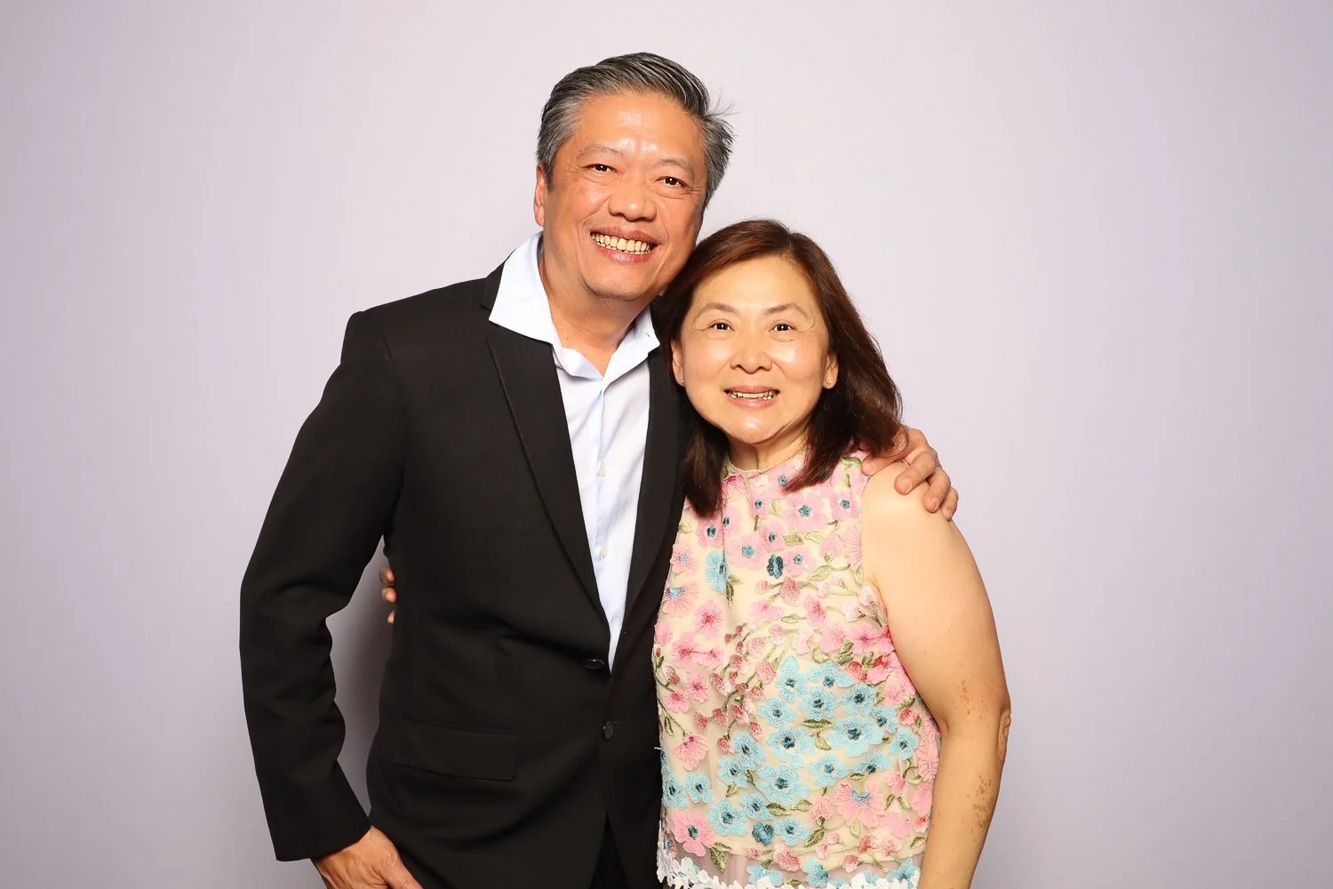 A middle-aged man and woman smiling and posing together in front of a light gray background. The man is wearing a black suit with a white shirt, and the woman is wearing a pink floral sleeveless top. The man's arm is around the woman's shoulder.