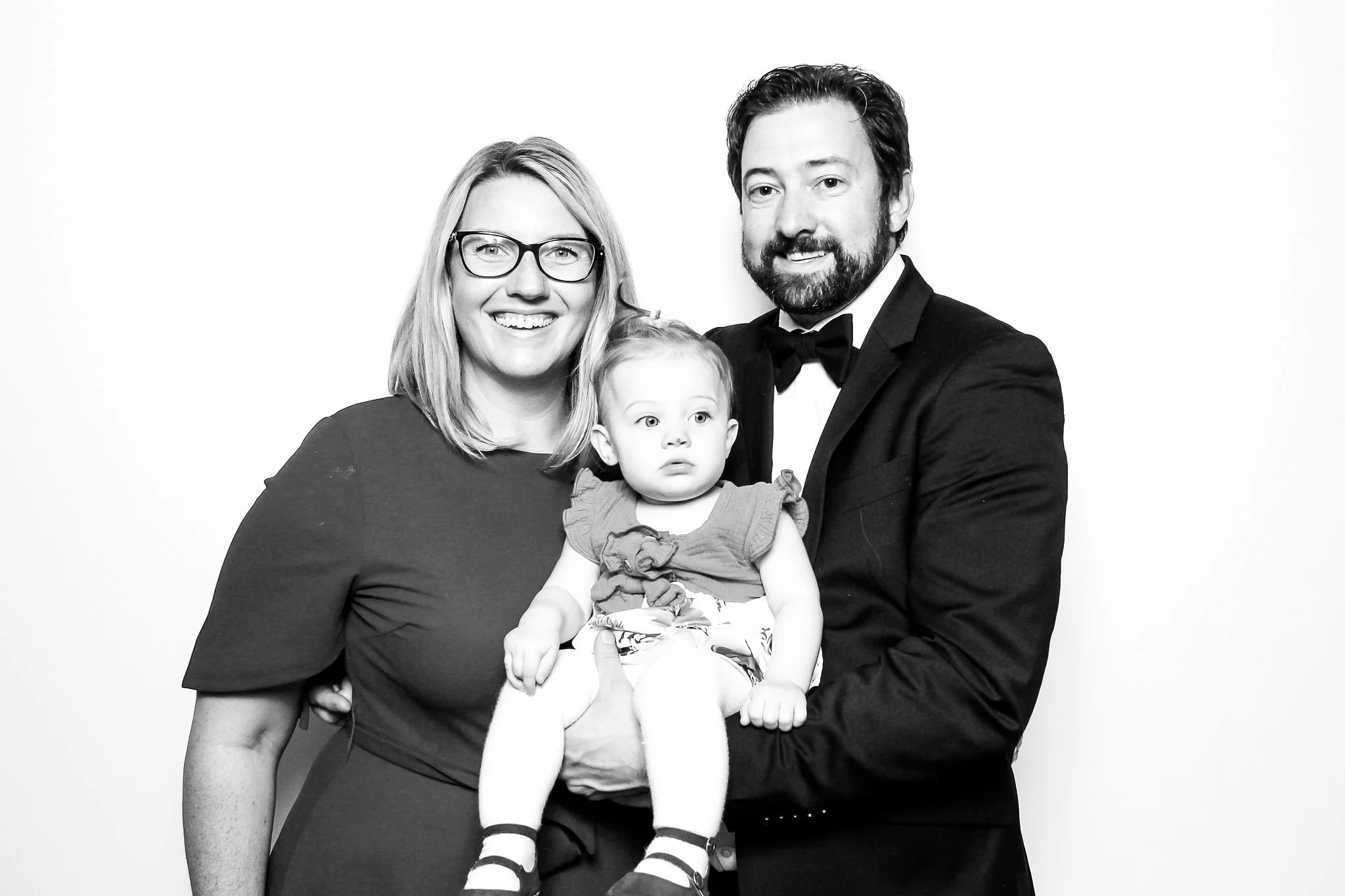 A happy family of three posing for a portrait, with a woman wearing glasses, a man in a tuxedo, and a young girl being held by the man against a plain white background.