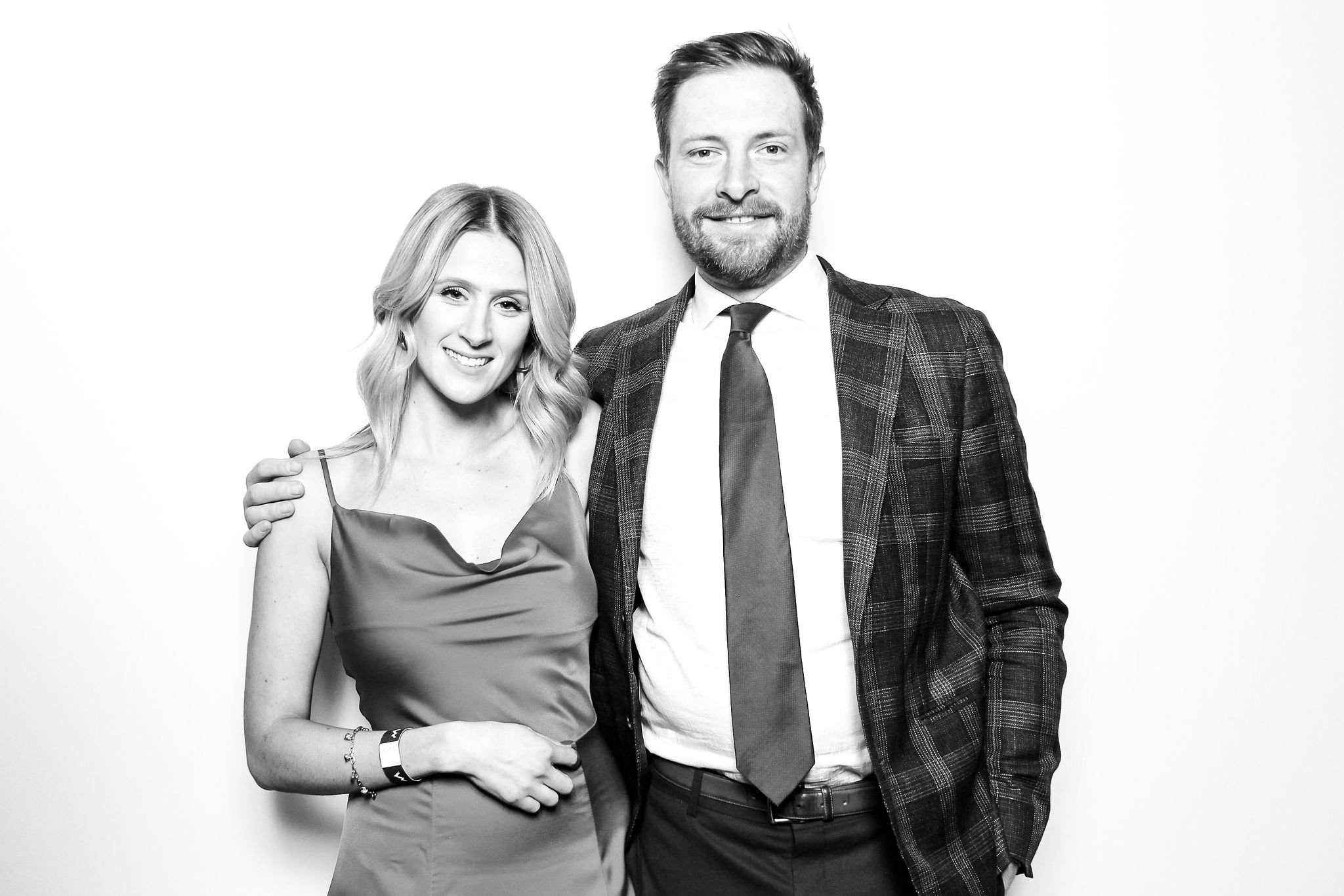 A black and white photo of a smiling man in a suit with a beard and a woman with wavy hair in a satin dress, standing together against a plain background.