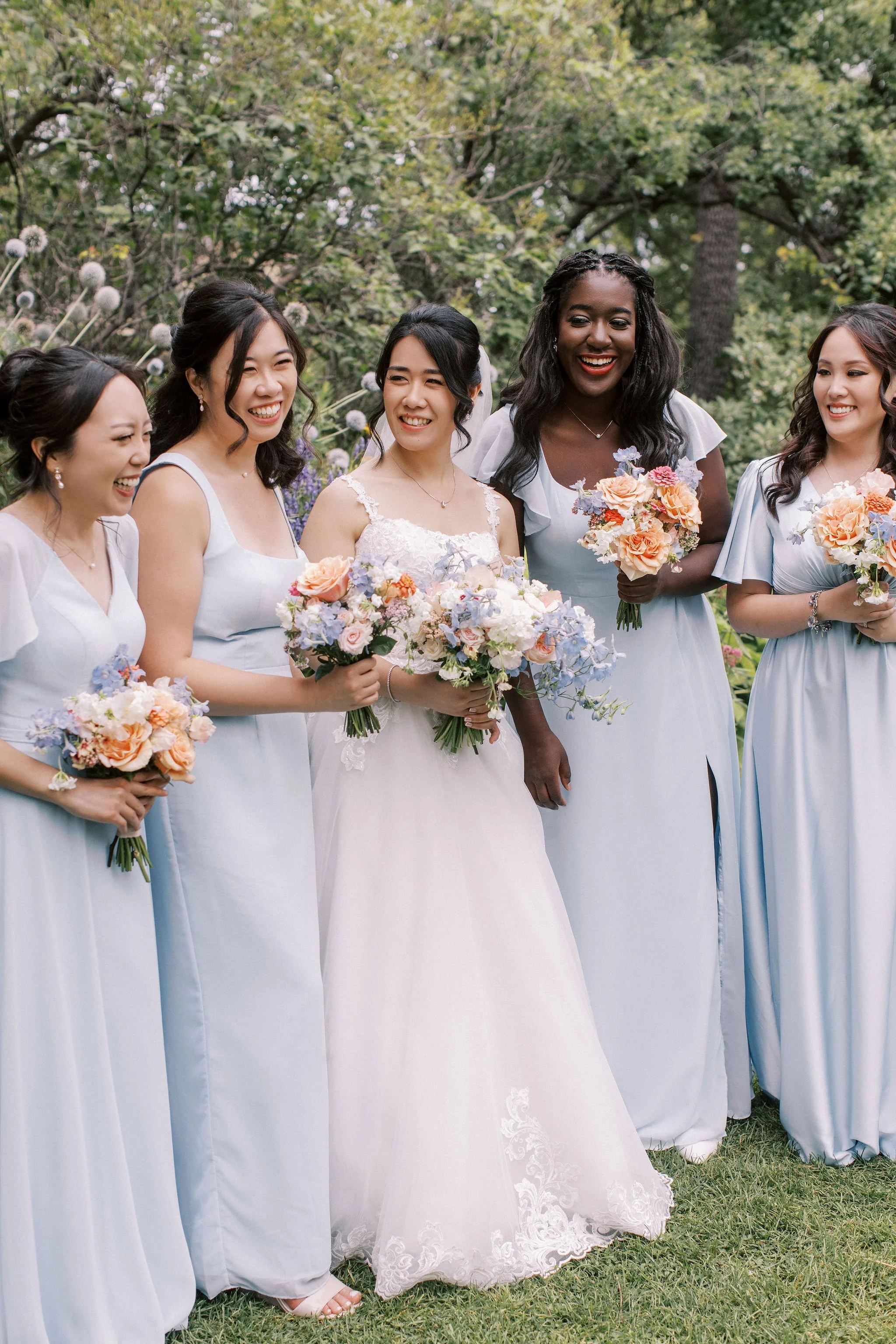 A group of bridesmaids and a bride standing outdoors, holding bouquets, smiling and celebrating during a wedding.