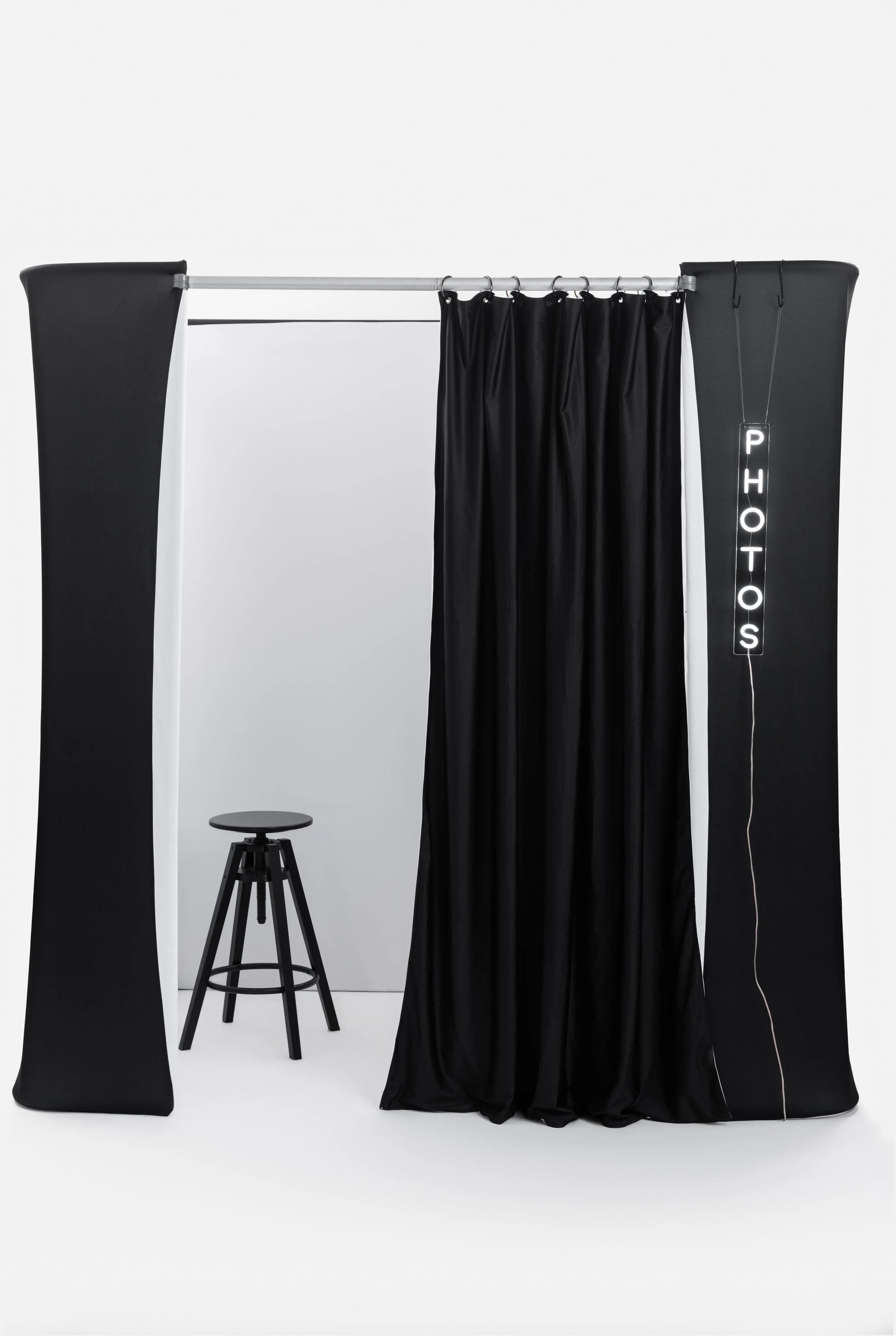 Photo booth with black curtains, a black stool, and a neon sign spelling 'PHOTOS' hanging on the right side.