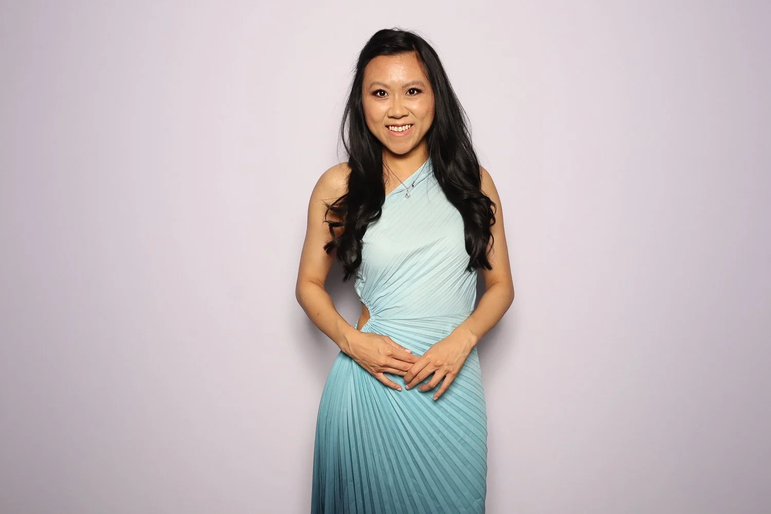 A woman with long, dark hair sits in front of a plain, light-colored background, smiling at the camera, wearing a sleeveless, pleated, light blue dress.
