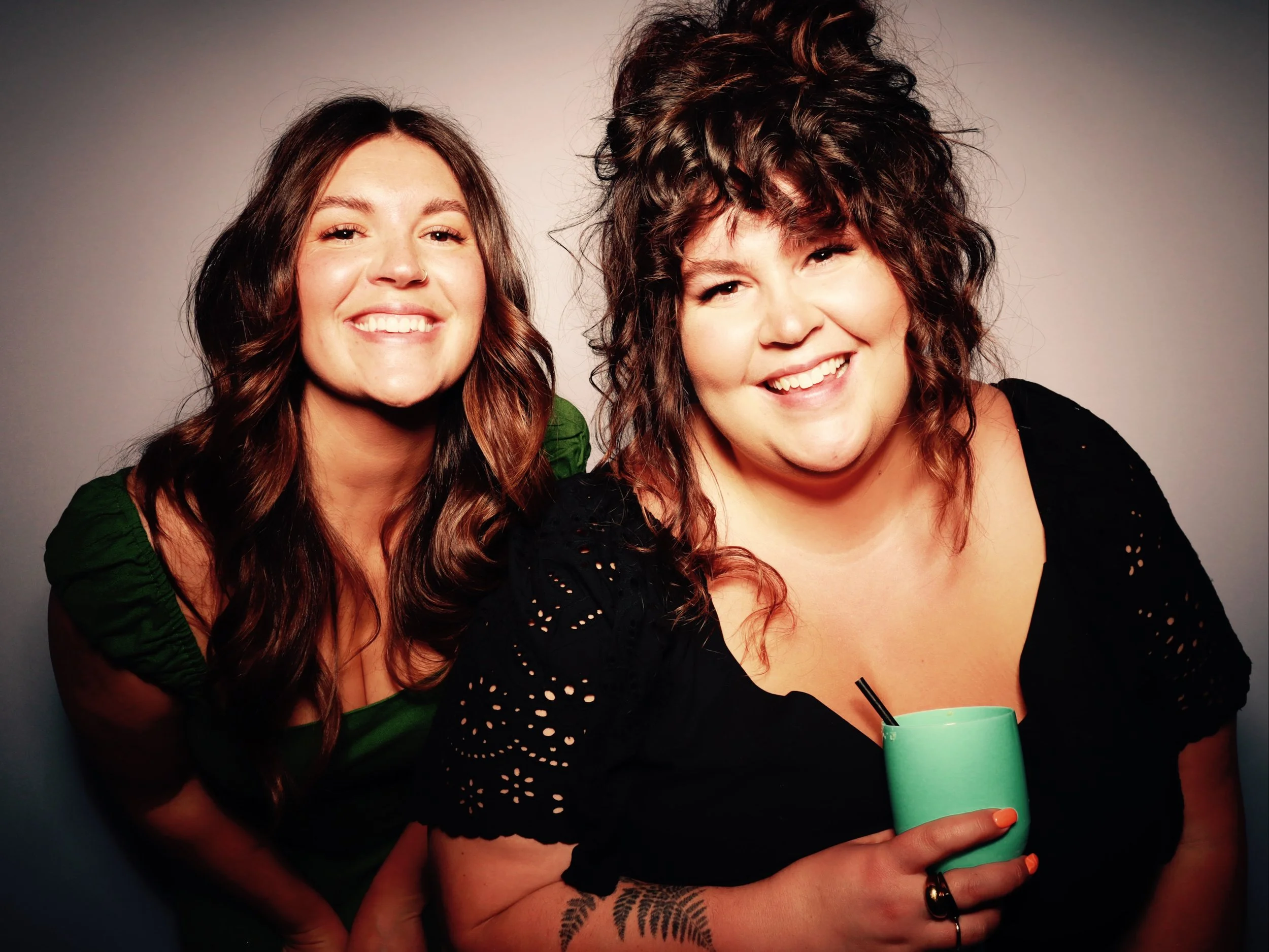 Two women smiling; one woman has long brown hair and is wearing a green shirt, the other has curly brown hair and is holding a teal cup with a straw, wearing a black top.