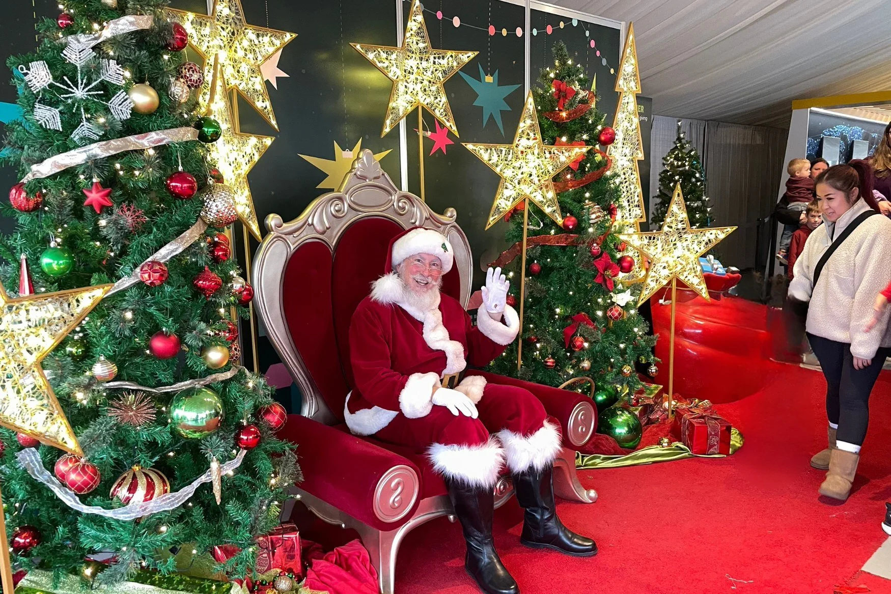 Santa Claus sitting on a red and silver throne, waving and smiling, surrounded by decorated Christmas trees with red, gold, and green ornaments, large illuminated star decorations, and wrapped Christmas presents on a red carpet. Children are standing nearby, one woman is smiling.