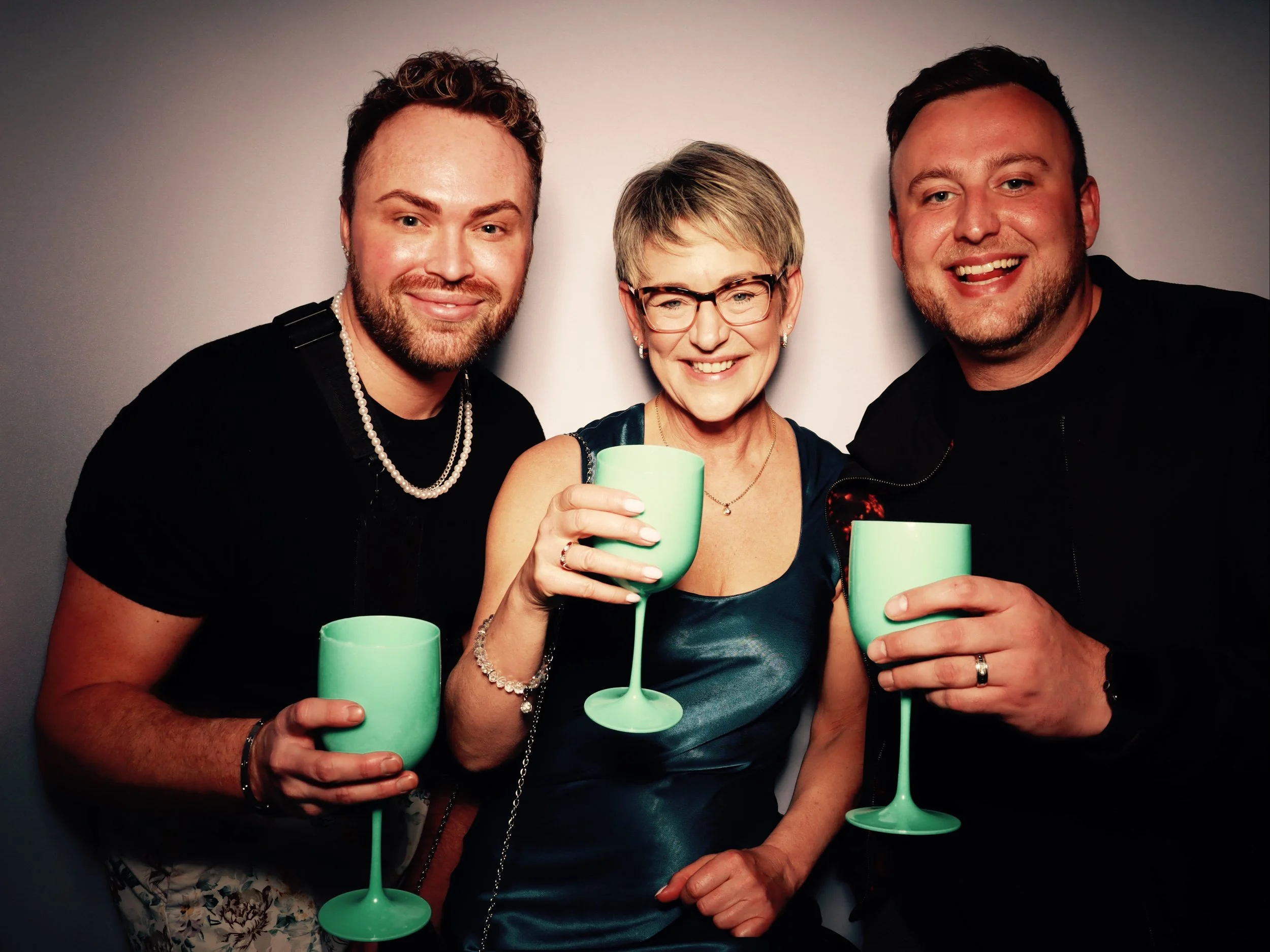 Three people smiling and holding green champagne glasses at a social gathering, posing for a photo in front of a plain background.
