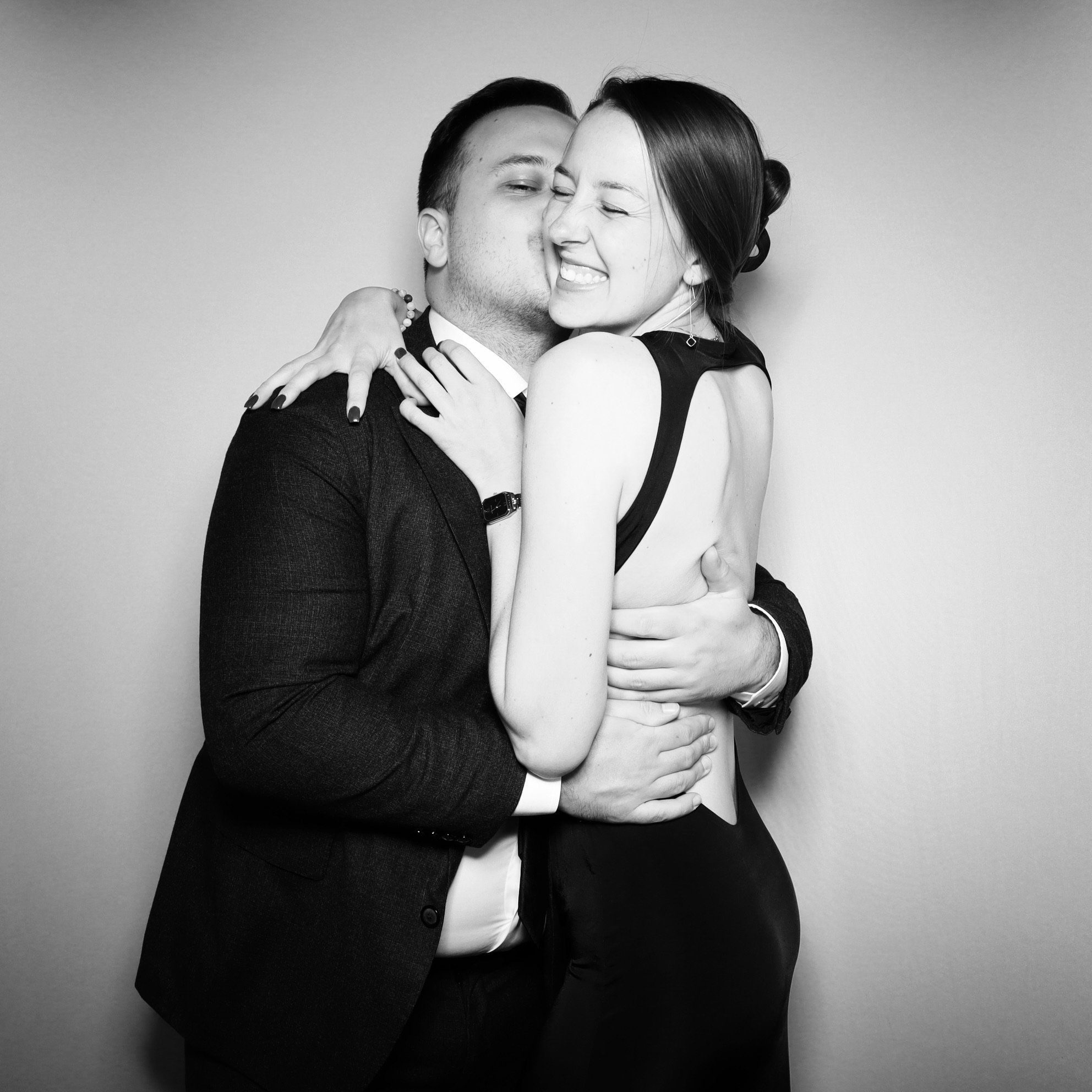 A black and white photo of a couple hugging and smiling, with the woman showing her teeth and eyes closed, the man kissing her on the cheek, both dressed formally