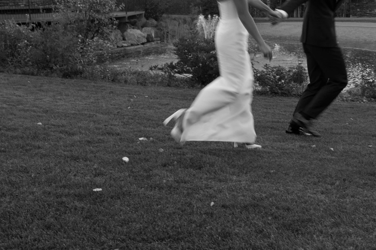 A bride and groom holding hands and dancing outdoors on a grassy area near a pond, dressed in wedding attire.