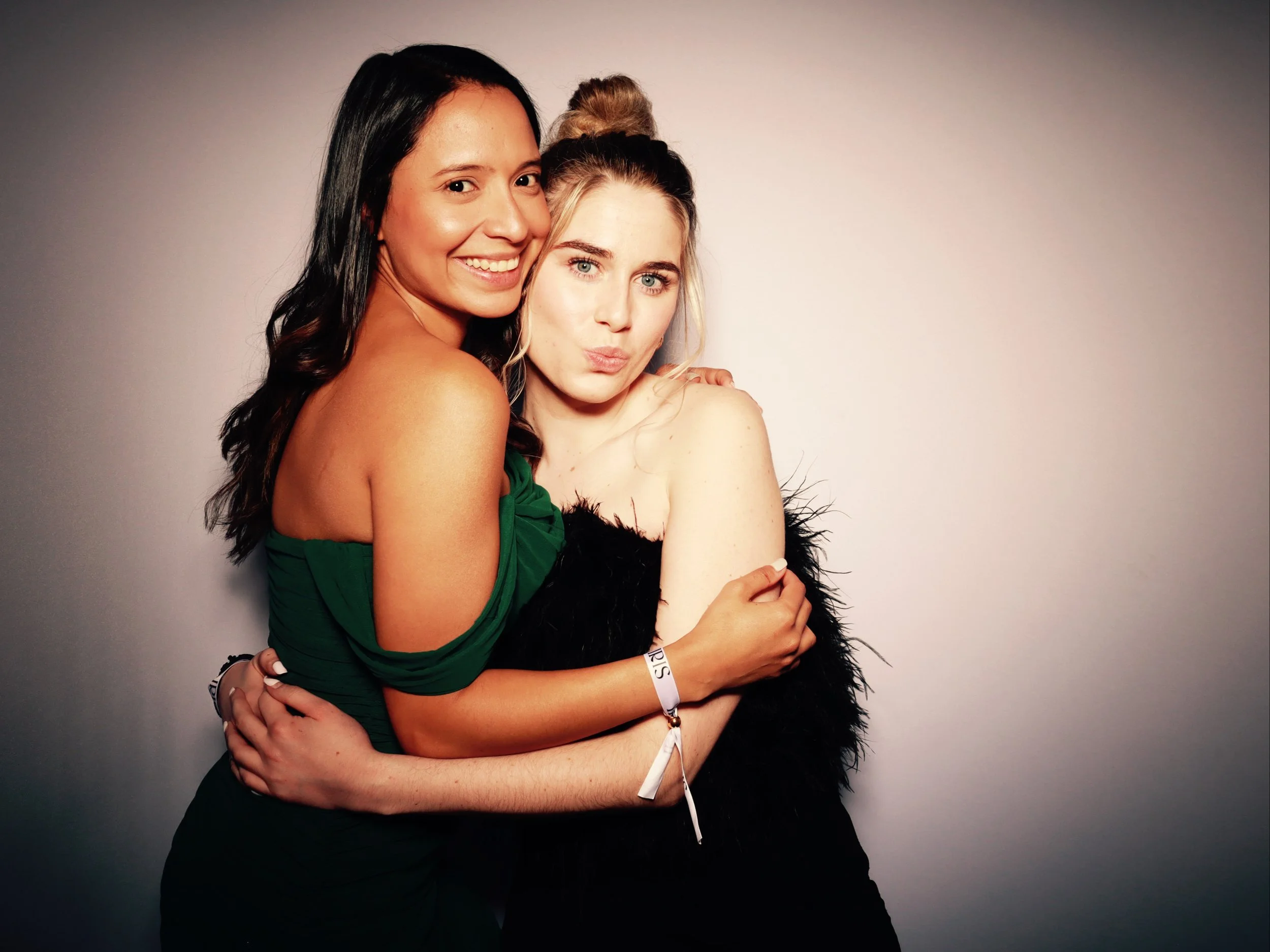 Two young women hugging and posing for a photo against a plain background.