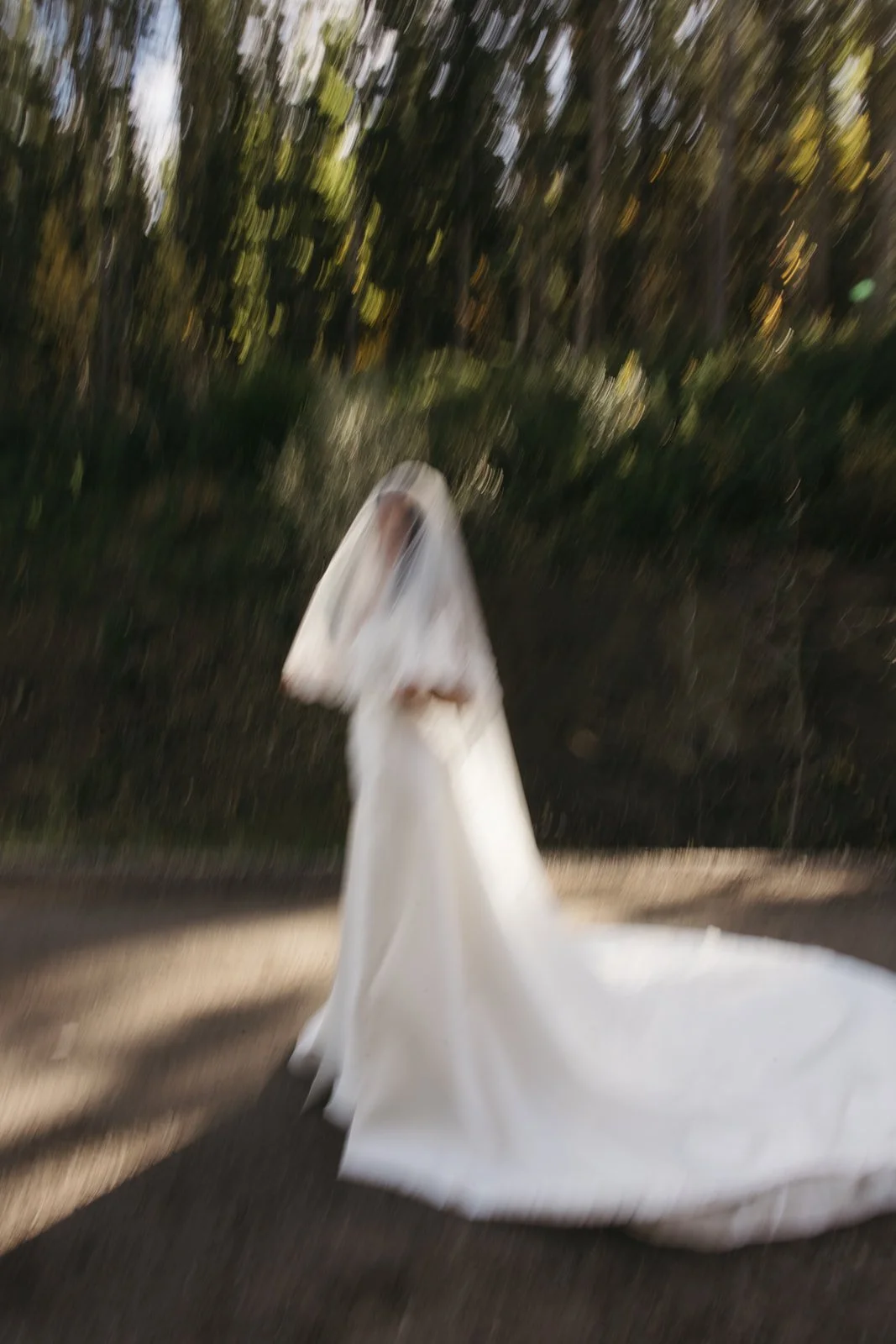 A person in a white dress and veil, possibly a bride, walking outdoors in a natural setting with trees and sunlight, with a motion blur effect.