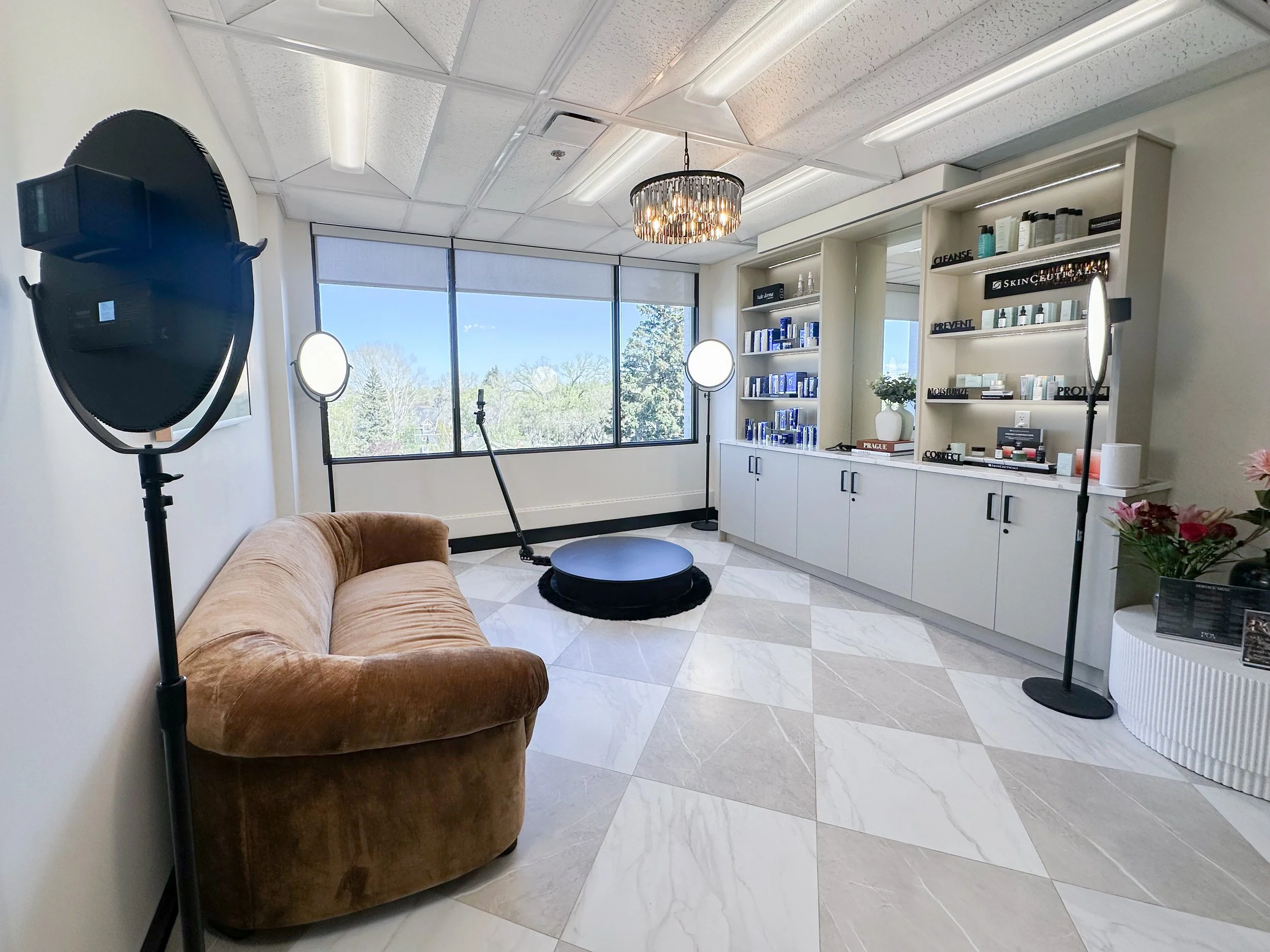 Interior of a spa or skincare clinic with a brown velvet sofa, large Windows with trees outside, shelves with skincare products, two round ring lights, a professional softbox light, a round platform for treatments, floral decorations, and a modern ch