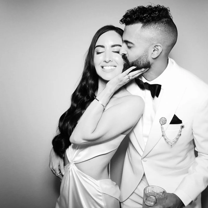 A couple at a party, with the woman smiling and the man kissing her on the cheek.