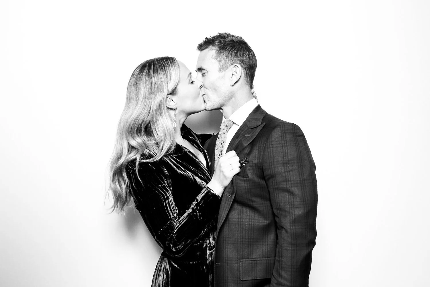A man and woman in formal attire kissing against a plain background.