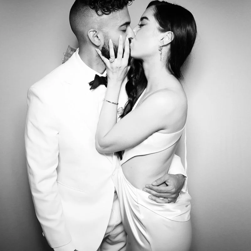 A black and white photo of a couple kissing, with the woman holding the man's face and the man wearing a white tuxedo with a bow tie.