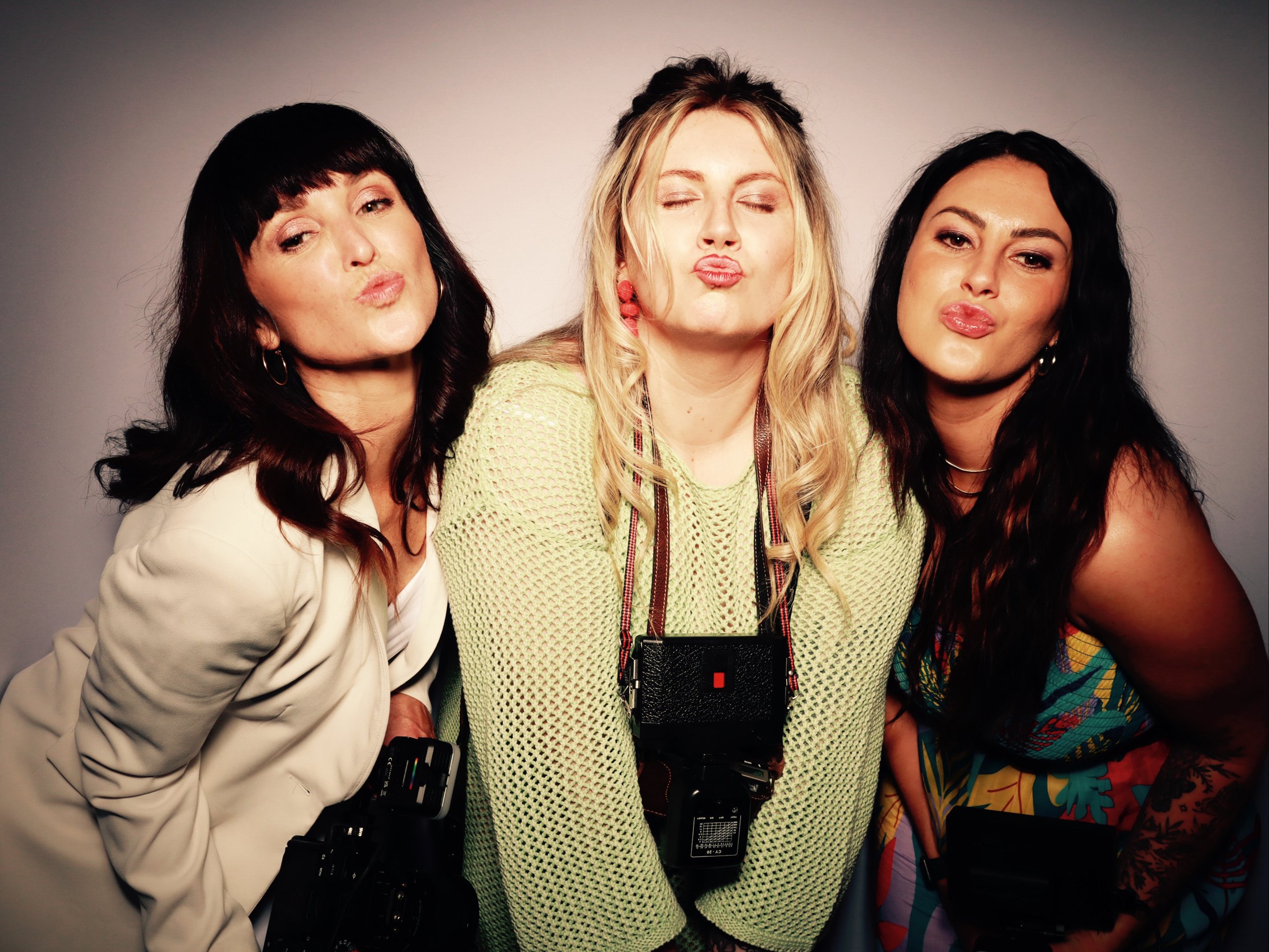 Three women posing together, making duck faces, with camera straps around their necks, standing against a plain background.