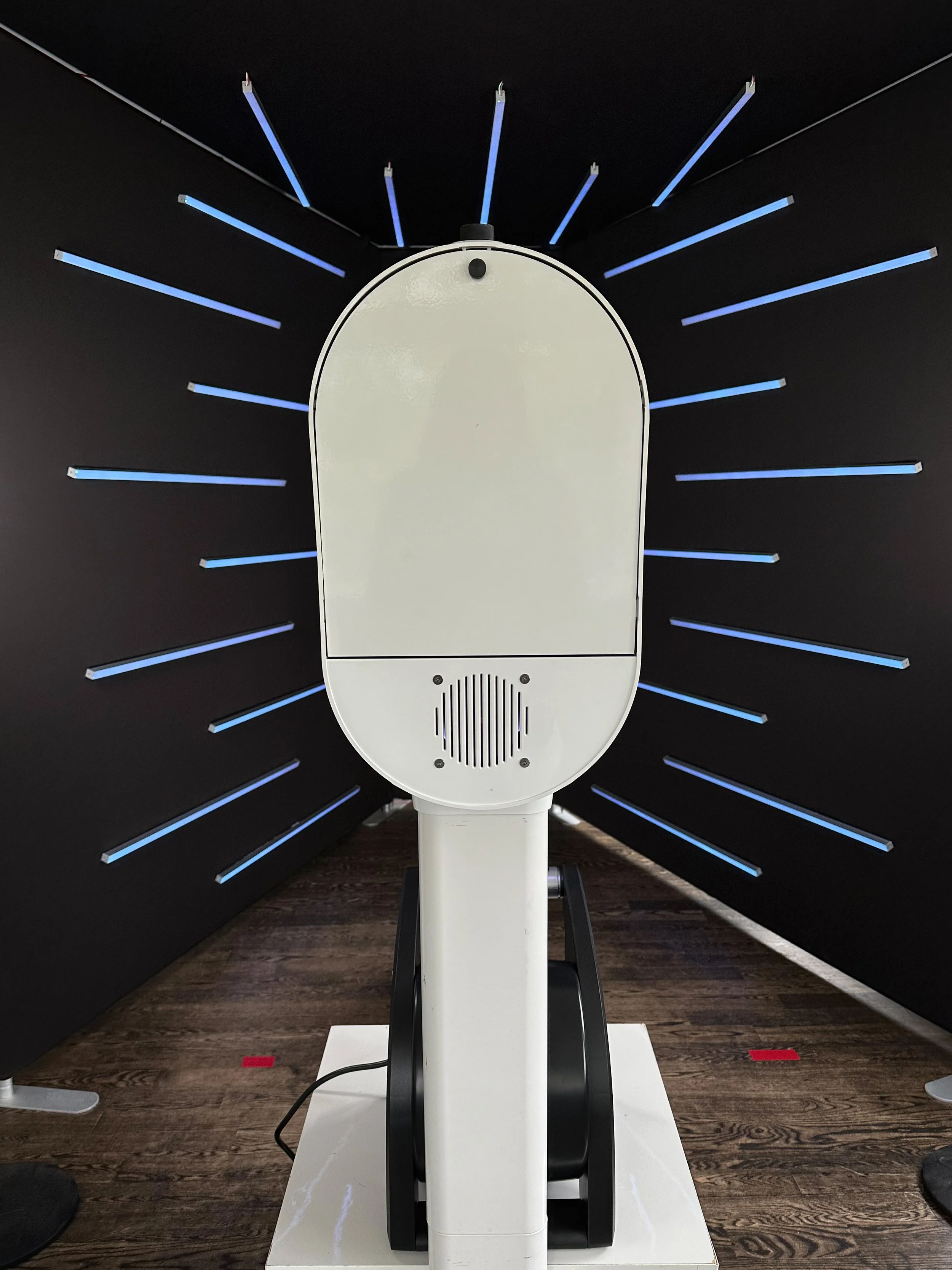 Front view of a white interactive kiosk or device with a rounded top, set against a black background with blue illuminated lines radiating outward.