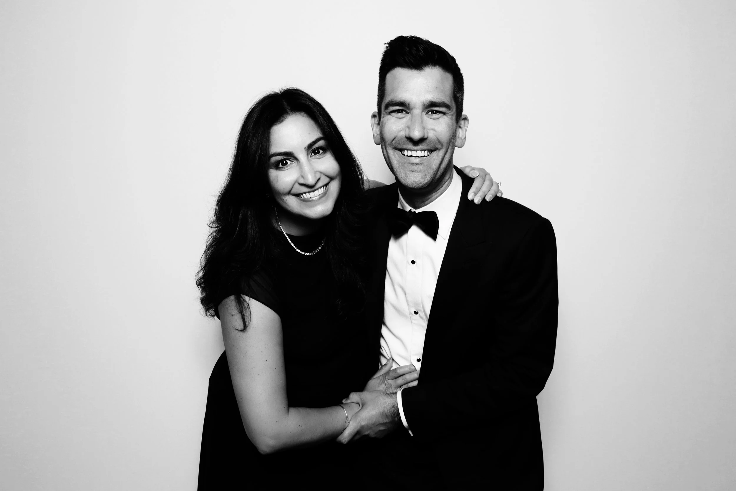 Black and white photo of a smiling man in a tuxedo and a woman in a black dress, hugging each other against a plain background.