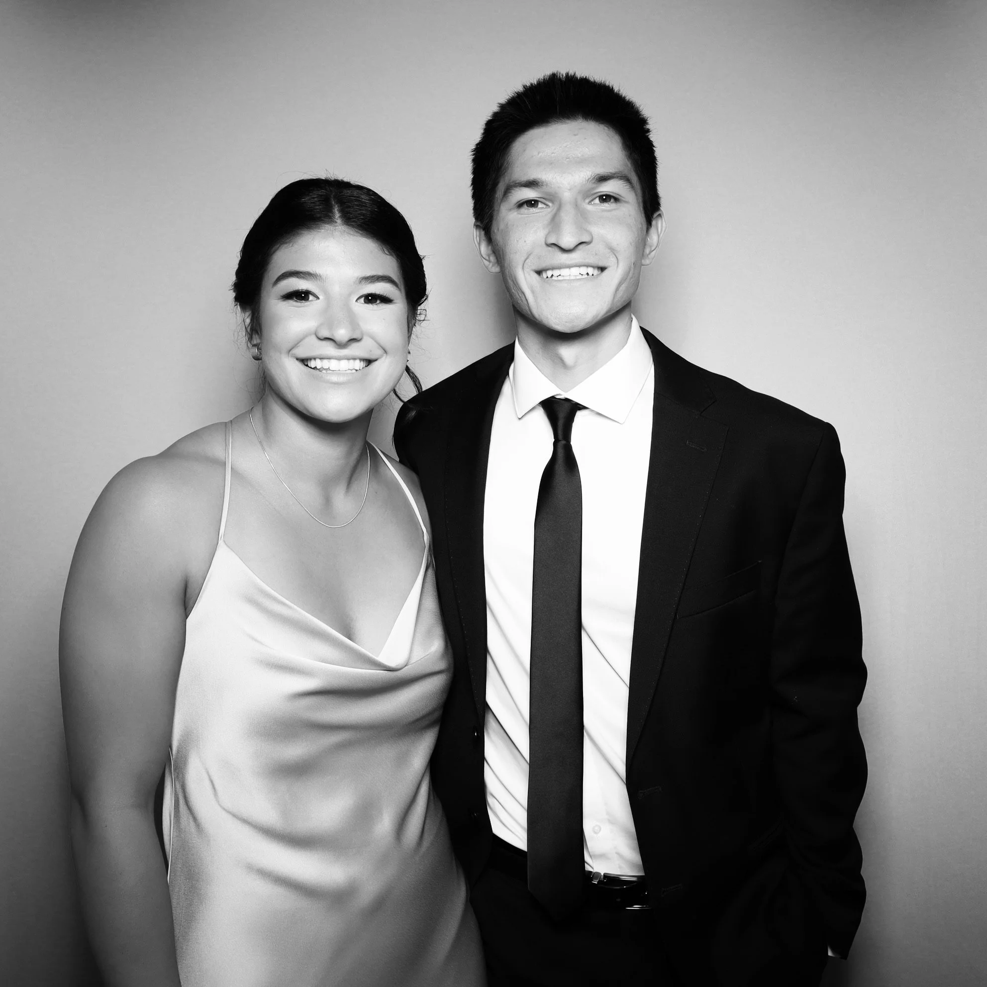 Black and white photo of a smiling young woman in a satin dress and a young man in a suit and tie posing together against a plain background.