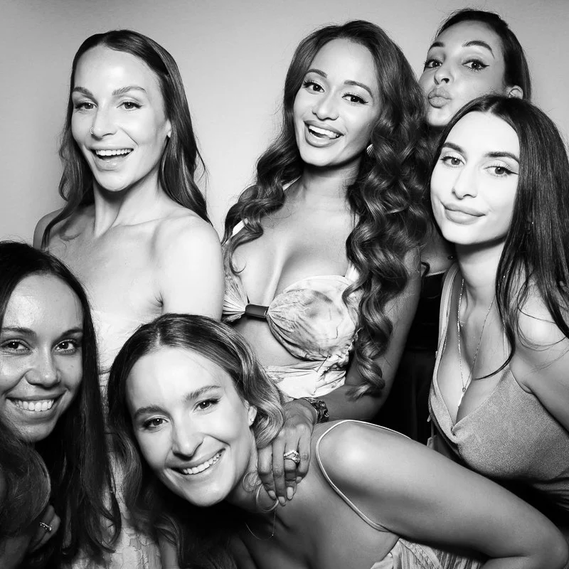 Six women smiling and posing together for a group photo, some making playful expressions, in a black and white image.