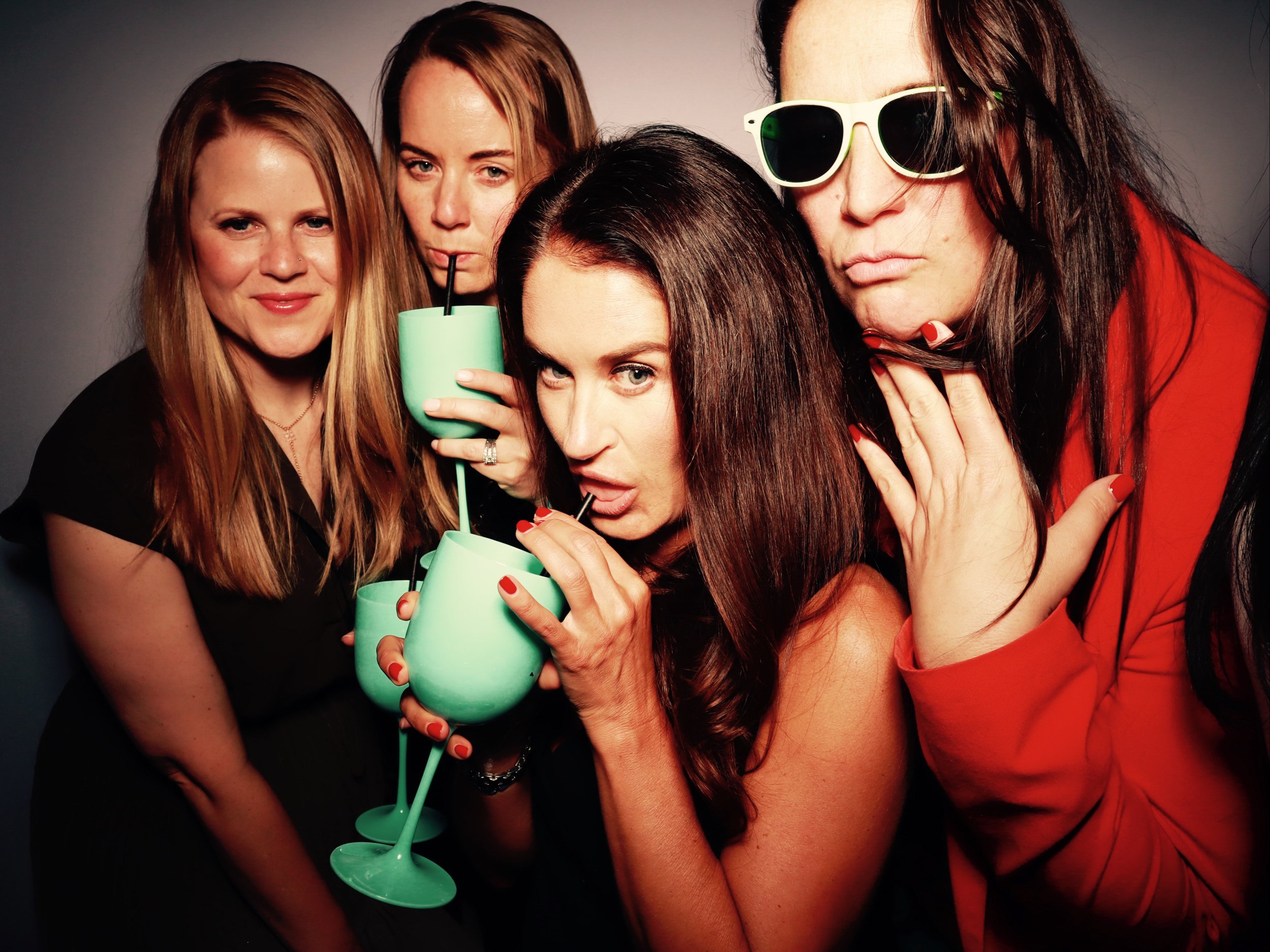 Four women at a party, holding green cocktails with straws, posing playfully for the camera. One woman is wearing sunglasses and a red jacket.