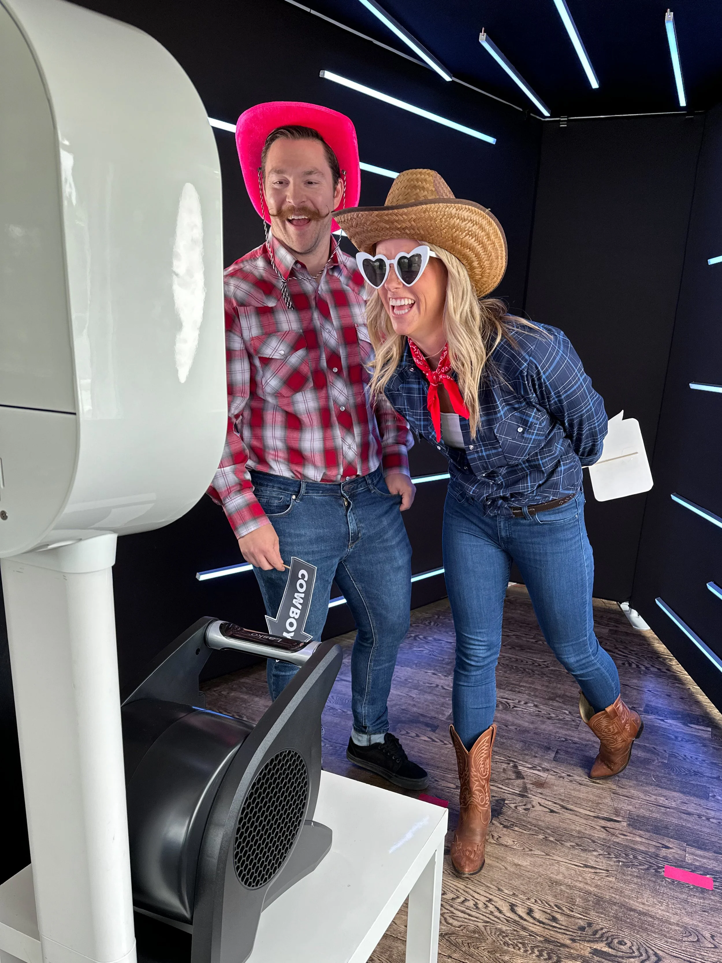 Two people dressed in cowboy-themed attire, wearing cowboy hats, bandanas, and western boots, standing next to a photo booth with fun expressions.