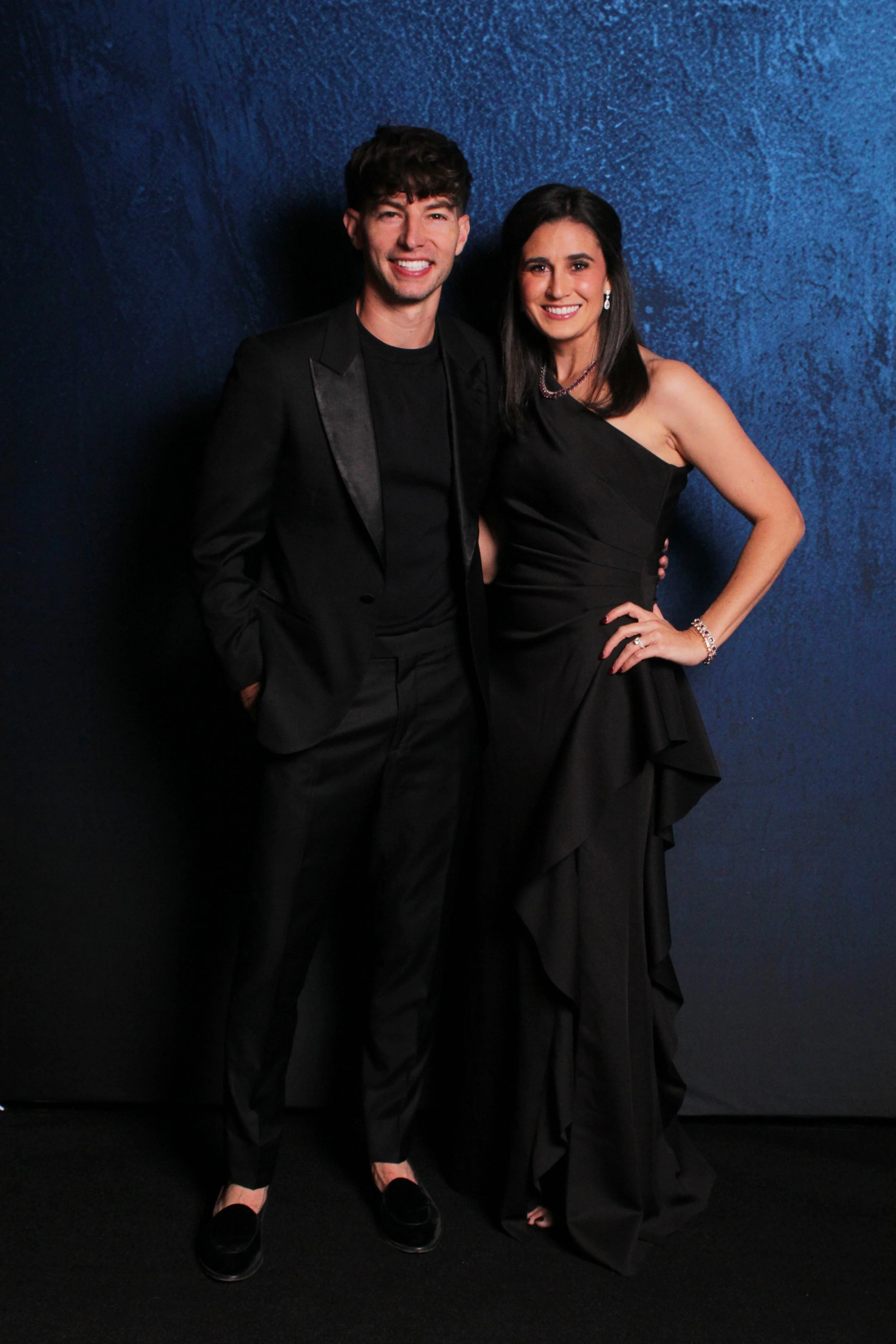 A man and woman in formal attire posing together against a textured blue wall.