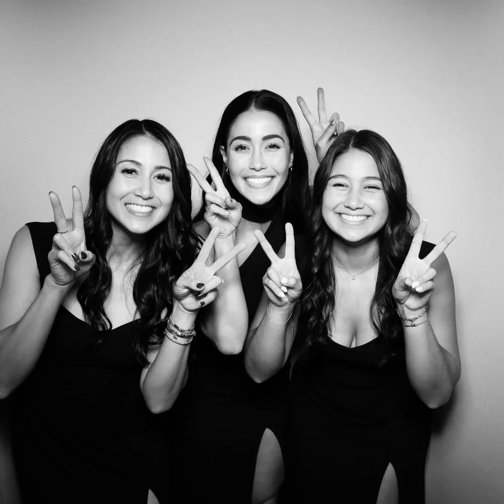 Three young women smiling and making peace signs with their fingers in a black-and-white photo.