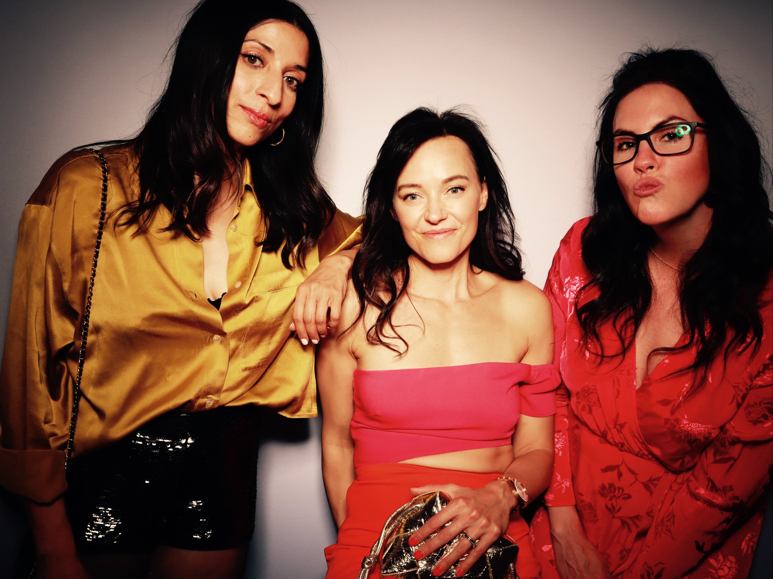 Three women posing closely together against a plain background, dressed in stylish clothes, with one woman in the center wearing a strapless pink top, the woman on the left in a gold blouse, and the woman on the right in a red floral dress.
