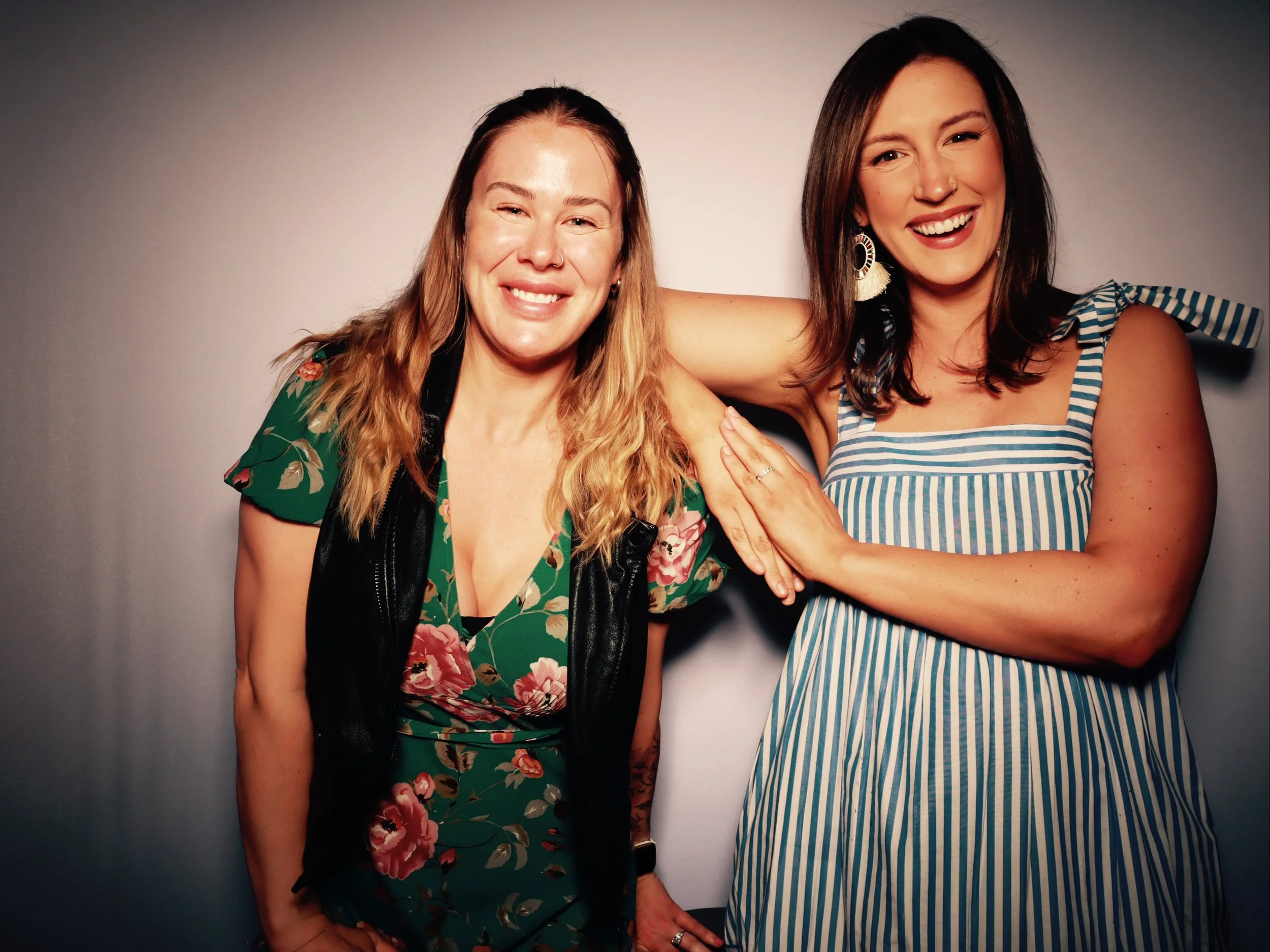 Two women smiling and posing together in front of a gray background. One woman has long blonde hair, is wearing a green floral dress with a black vest, and is squinting. The other woman has shoulder-length dark hair, is wearing a blue and white strip