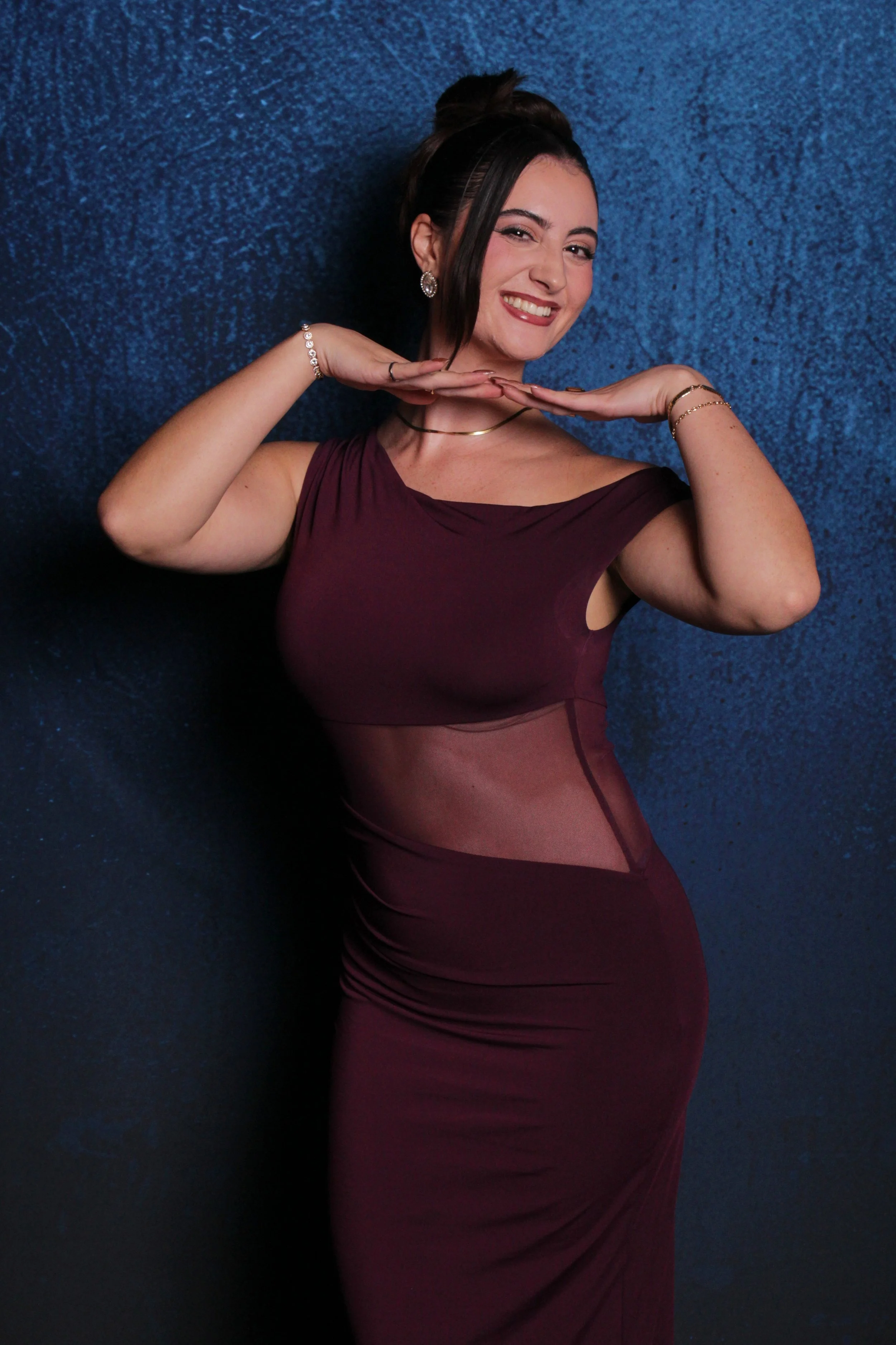 A woman in a burgundy dress with sheer side panels is posing against a blue textured background. She is smiling, with her hands framing her face and head slightly tilted, wearing jewelry and her hair styled in an updo.